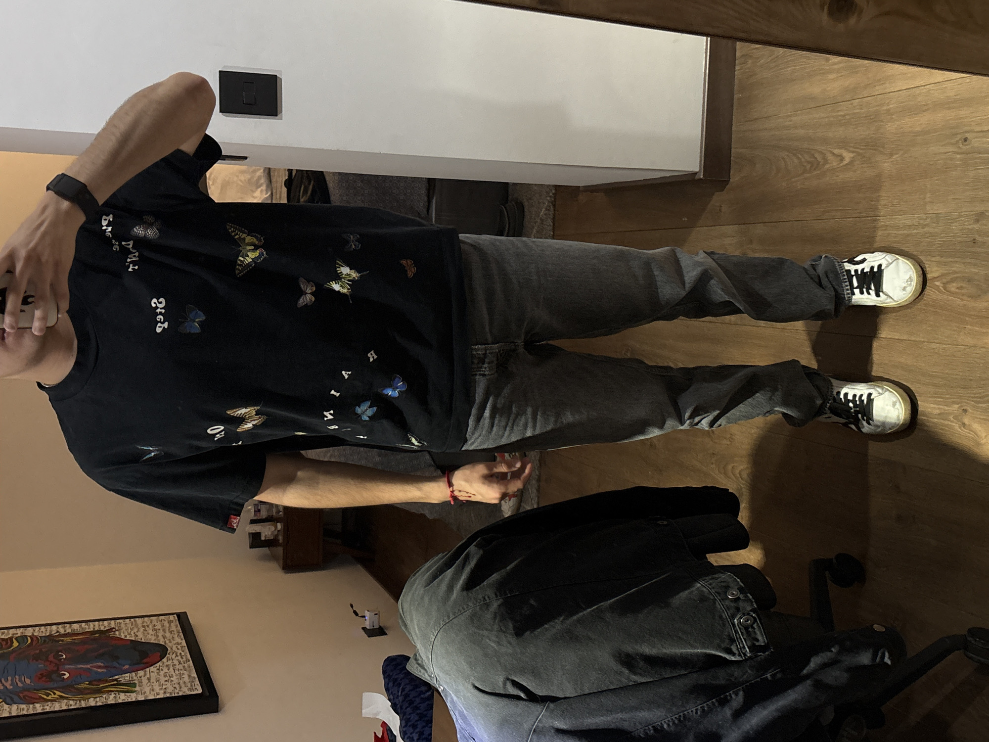 A person stands on a wooden floor, wearing a black t-shirt with butterfly designs and gray jeans. The person is also wearing white sneakers with black laces and has a black smartwatch on their left wrist. The person's left hand is near their face, possibly touching or adjusting something. To the right, the floor transitions into a different surface. A black jacket rests on a chair. The background shows a white wall with a black switch, a framed artwork on a wall, and a glimpse of other furniture. The location appears to be inside a home, and there is no definitive information to confirm the city of San Bernardino Tlaxcalancingo, Mexico.