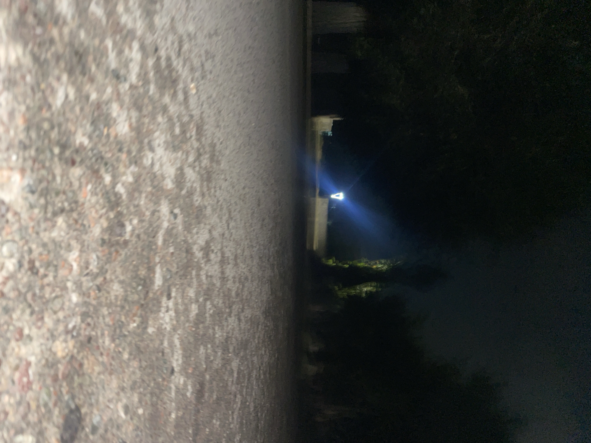The image depicts a dark, outdoor scene, likely at night, on a paved or gravel surface in Ak-Jol, Kyrgyzstan. The foreground is filled with a blurred texture of small stones and pebbles, suggesting a pathway or ground cover. In the midground, a bright, triangular light source emanates a blue-white glow, casting illumination across the path and creating a lens flare effect. This light appears to be from a streetlamp or similar fixture, positioned in front of some dark, indistinct structures and trees that line the background. The overall environment is dimly lit, with the darkness dominating the scene, creating a sense of quiet and isolation. No individuals or specific actions are discernible in the image due to the low light conditions and the focus on the overall atmosphere of the location.