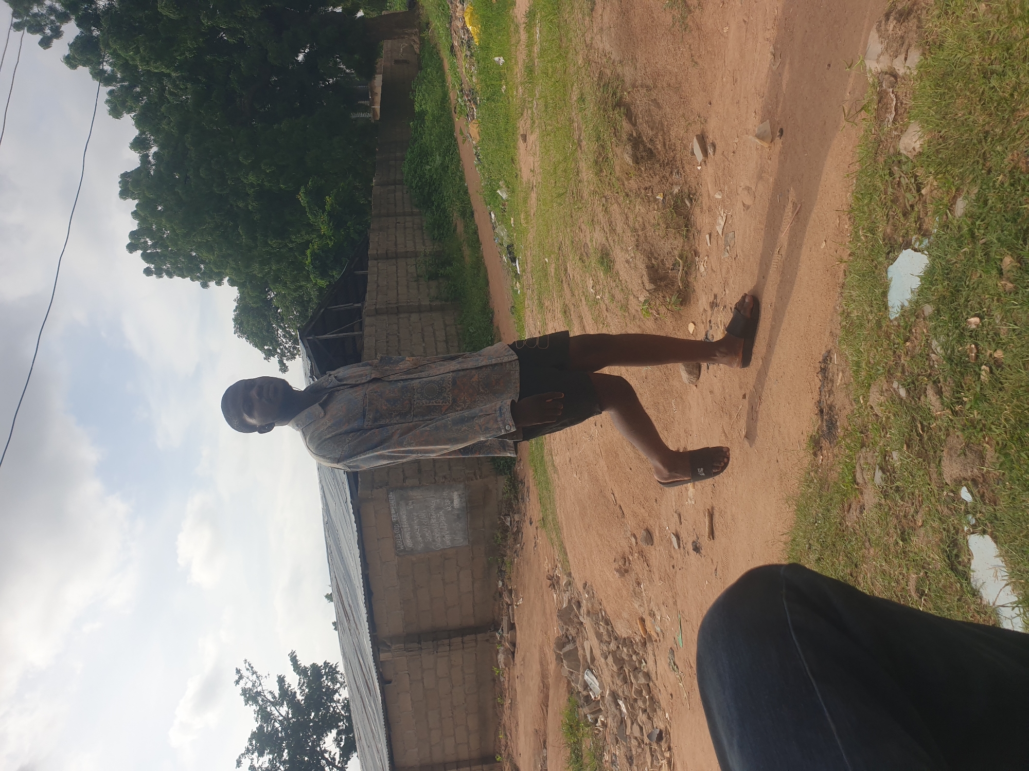 A young boy, standing in a dirt path, is captured in a photograph. He is wearing a patterned shirt and dark shorts, with sandals on his feet. The path is lined with grass and scattered debris, suggesting an outdoor, possibly rural, setting. In the background, a brick building with a corrugated metal roof is visible, along with a large tree. The sky is cloudy, indicating it might be daytime. There is some writing on a dark surface on the wall of the building, but it is too blurry to read. The overall impression is that of a casual snapshot taken in a humble environment.