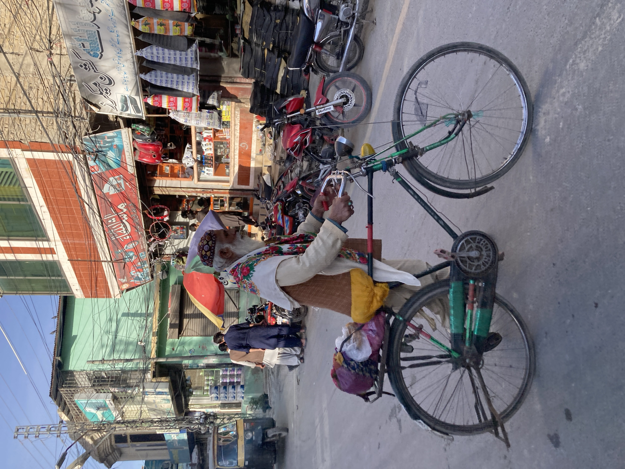 An elderly man with a long white beard and a patterned skullcap is riding an old bicycle down a street in Quetta City Tehsil, Pakistan. He is wearing a light-colored shirt and vest, with a colorful scarf around his neck. The bicycle has green accents, and various items are tied to it, including bags and what appears to be a red container covered with yellow fabric.
The street is lined with shops, some displaying goods such as clothing, tires, and auto parts. There are motorcycles parked along the street. Several people are walking in the background. Electrical wires crisscross overhead. The scene suggests a busy, possibly older commercial area within the city.