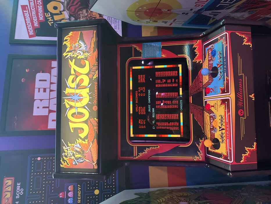 The image showcases a vintage "Joust" arcade machine with vibrant artwork on its sides. The screen displays high scores from "Daily Buzzards" and "Joust Champions," listing player initials and scores. The control panel features two sets of joysticks and buttons for two-player action. The surrounding area includes framed posters of "Red Dawn," a Pac-Man machine, and another partially visible poster, creating a retro arcade ambiance.