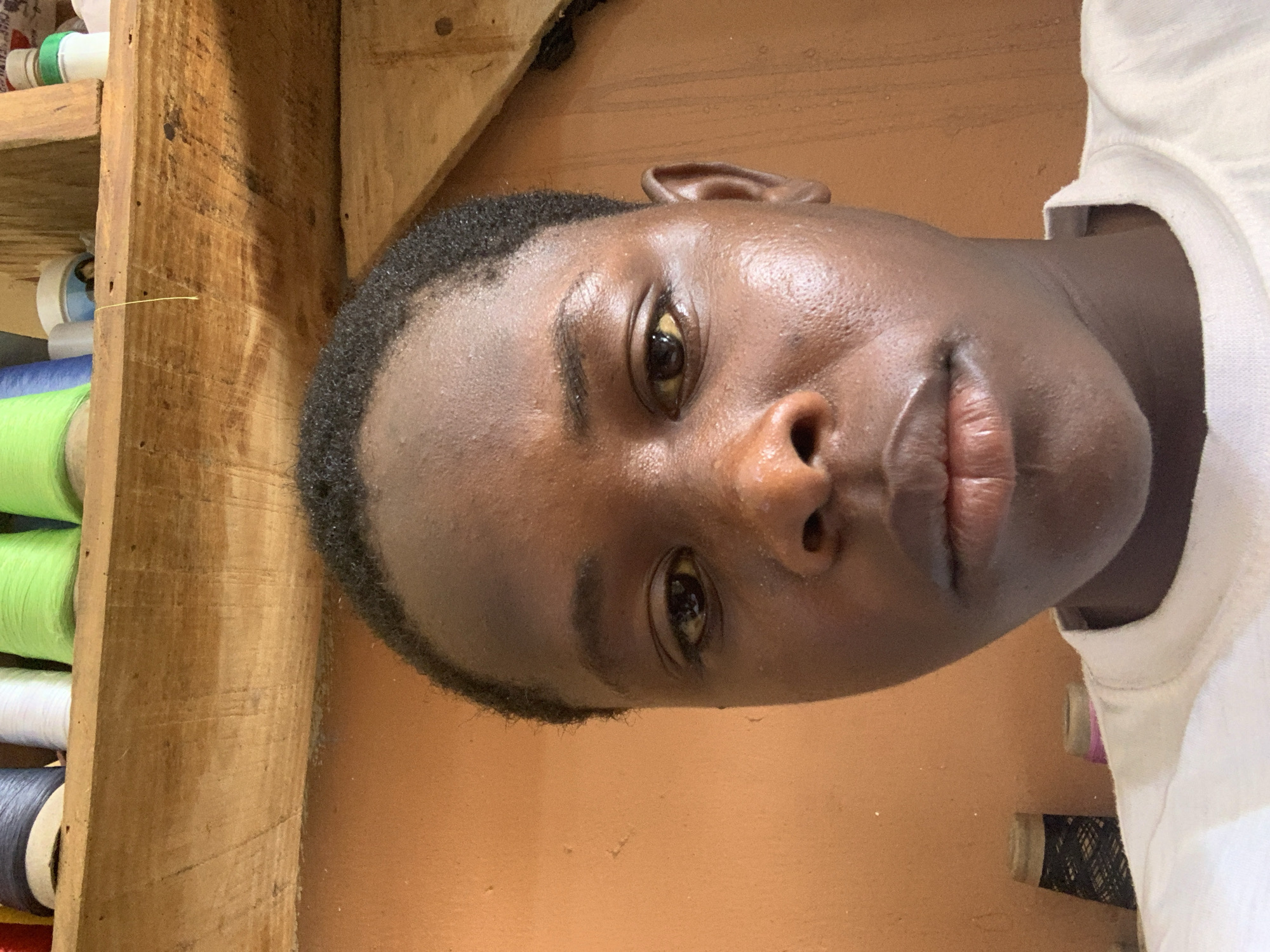 The image is a close-up portrait of a young Black individual's face, taken from a slightly low angle. The subject has dark skin, short, dark, curly hair, and is wearing a white t-shirt. Their expression is neutral. The background consists of an orange-colored wall and a wooden shelf above, on which several spools of colorful thread are visible, suggesting a craft or sewing environment. The lighting is somewhat harsh, highlighting the texture of the skin. The image does not provide any specific visual cues to pinpoint its location within the city of Jos, Nigeria.