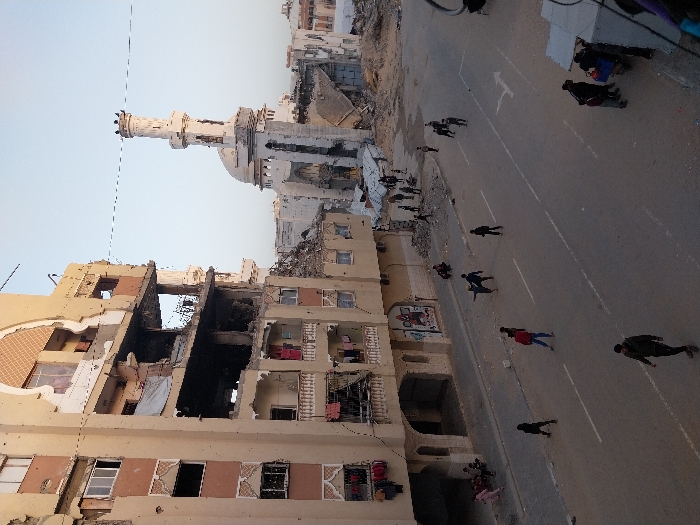 The image depicts a scene of extensive urban destruction, likely in the aftermath of a conflict or disaster. The photo appears to be taken from a slightly elevated vantage point, looking down a street lined with heavily damaged buildings.
In the foreground to the left, a multi-story building, likely residential or commercial, shows severe structural damage. Large sections of its walls are missing, exposing the interiors of several floors. Darkened areas suggest fire damage or debris. The building's facade is light brown and beige, with some decorative tiling visible on lower levels. What appears to be laundry or fabric hangs from exposed windows or balconies, indicating that some areas might still be inhabited or recently vacated.
Across the street, another building on the right also exhibits damage, with rubble and exposed structures. Further down the street, a prominent mosque with a tall, light-colored minaret and a dome is visible. The mosque itself appears to have sustained damage, with visible wear and possibly missing architectural elements. Beyond the mosque, more buildings in the distance are seen as partially collapsed or reduced to rubble, suggesting widespread devastation. Piles of debris are scattered throughout the background.
The street itself is paved, likely asphalt, with white lane markings and a white arrow indicating traffic flow away from the viewer. Numerous people are scattered along the street, walking in various directions. Most individuals are dressed in dark clothing, though one person in the mid-ground wears a red top. Some people appear to be moving in small groups, while others walk alone. One individual closer to the mosque seems to be pushing a cart or carrying a large bundle. Their presence suggests ongoing activity and movement within the damaged environment.
The sky is a clear, light blue-gray, indicating daylight. The lighting suggests it might be late afternoon or early morning, with soft, diffused light and subtle shadows. The overall atmosphere conveyed is one of immense destruction but also of human presence and movement amidst the ruins. No clearly legible text is visible on any signs or structures in the image.