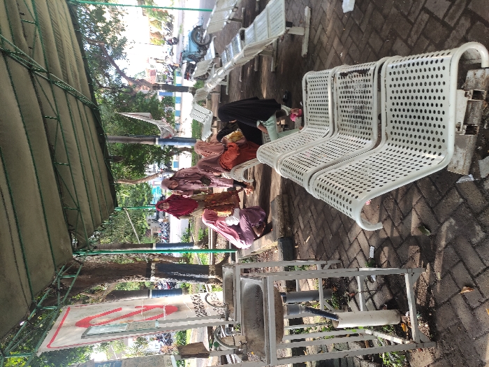 This outdoor image captures a public waiting area in Situbondo, Indonesia, under bright daylight on what appears to be a clear, sunny day. A green canvas canopy provides shade over a brick-paved ground.
The scene features several women, most of whom are wearing hijabs in various colors, including light brown, maroon, reddish-purple, black, and light purple/pink. They are seated on or near white, perforated metal benches, appearing to be resting or waiting. One woman in a light green top is partially visible behind others.
In the foreground, a utilitarian hand-washing station is prominent. It consists of a metal frame supporting a basin and is equipped with PVC pipes, suggesting a foot-operated water dispenser. Adjacent to this station, a white sign displays a red, stylized logo with faint, unreadable text below it. Another small, unreadable sign is visible nearby.
The white perforated metal benches, characteristic of public facilities, are arranged in rows, with some occupied and others empty. The background reveals trees and greenery, hinting at a natural environment beyond the immediate waiting zone. Distant motorbikes and a blue and white striped barrier suggest the presence of a nearby road or parking area. The overall atmosphere is calm and passive, typical of a public gathering or waiting point.