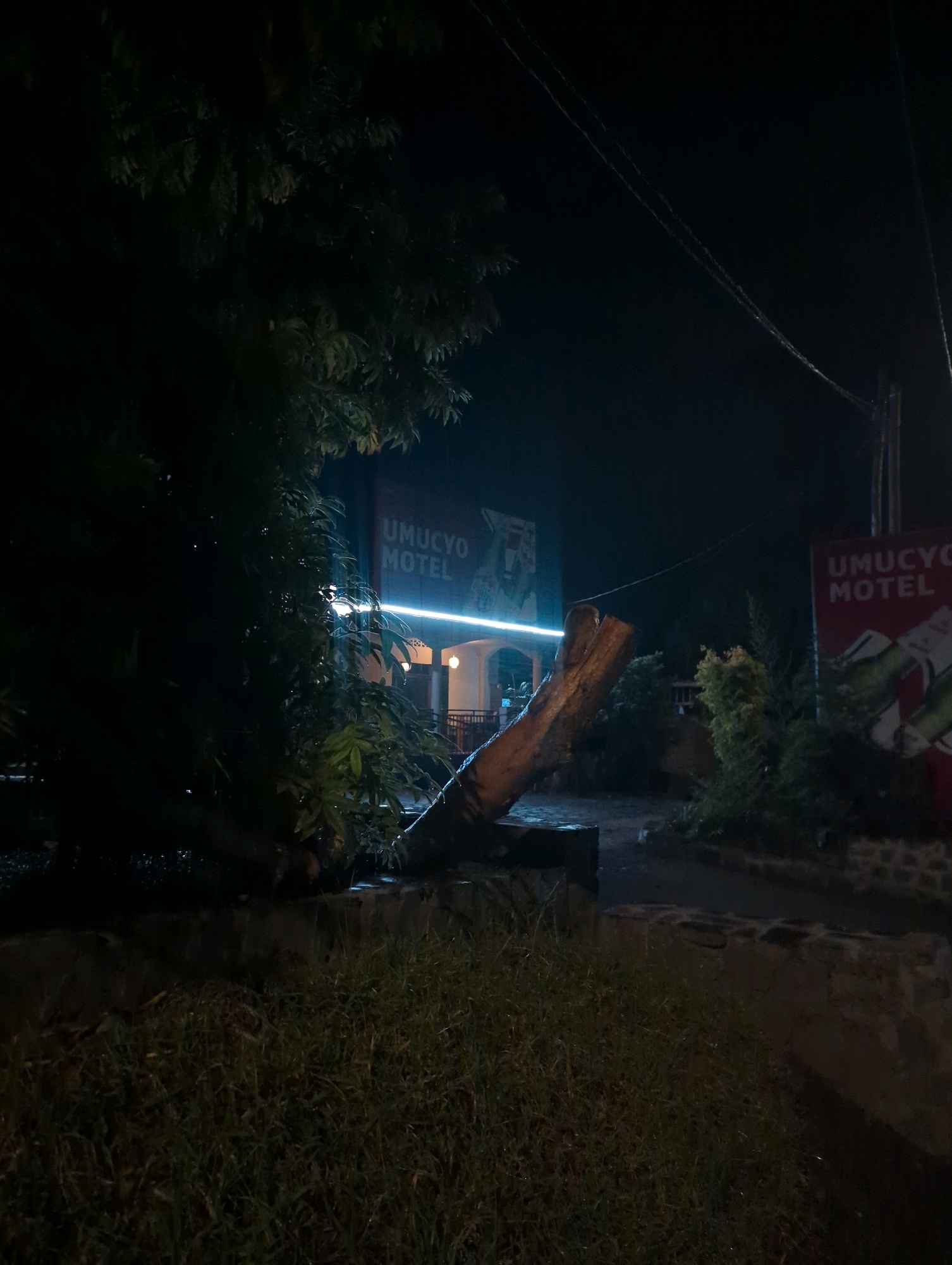 The image captures an exterior nighttime view of the UMUCMYO MOTEL in the Musanze District of Rwanda. A prominent, illuminated sign for "UMUCYO MOTEL" is visible on the left side of the frame, with a similar sign on the right. A large, rough-hewn log is positioned diagonally in the foreground, resting on a low stone structure. Trees with dark foliage frame the scene, and power lines are strung across the dark sky. The ground appears wet, suggesting recent rain. The entrance to the motel, with visible lighting, is in the mid-ground, and a railing encloses a walkway. The overall atmosphere is dimly lit and moody.