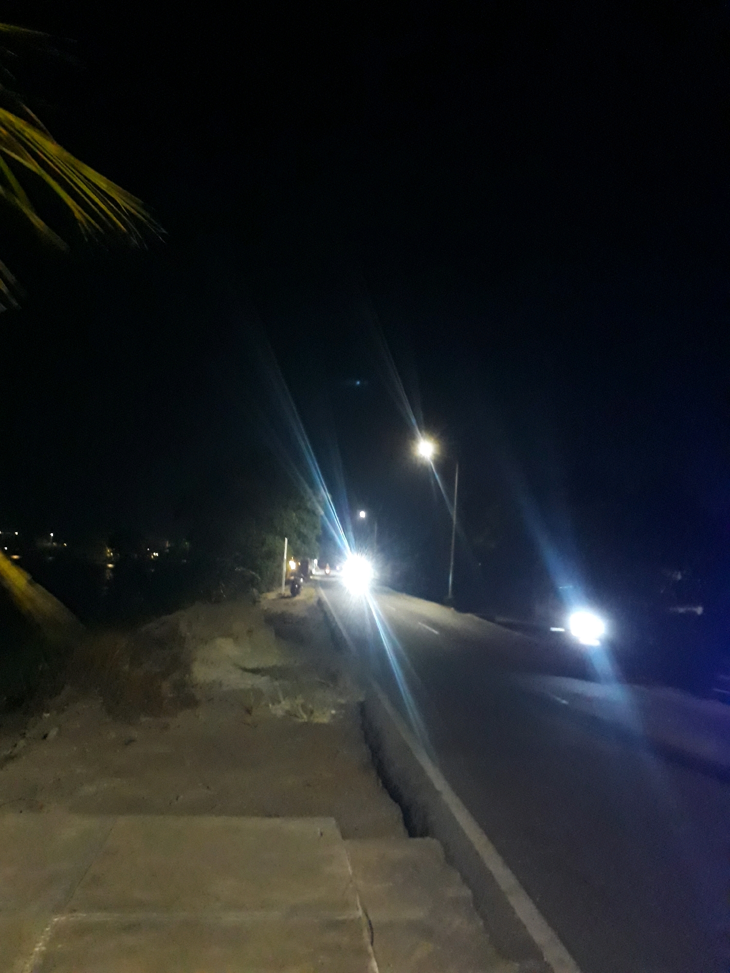 The image depicts a nighttime street scene in Pangkalpinang, Indonesia. A road stretches into the distance, illuminated by streetlights and the headlights of approaching vehicles. On the left side of the road, there is a sidewalk or pathway, partially obscured by a dark area and a palm frond in the upper left corner. The road itself appears to be asphalt and is bordered by a concrete curb. In the distance, several lights suggest buildings or other structures. The overall atmosphere is dark, with the artificial lights creating strong glare and lens flares due to the night conditions.