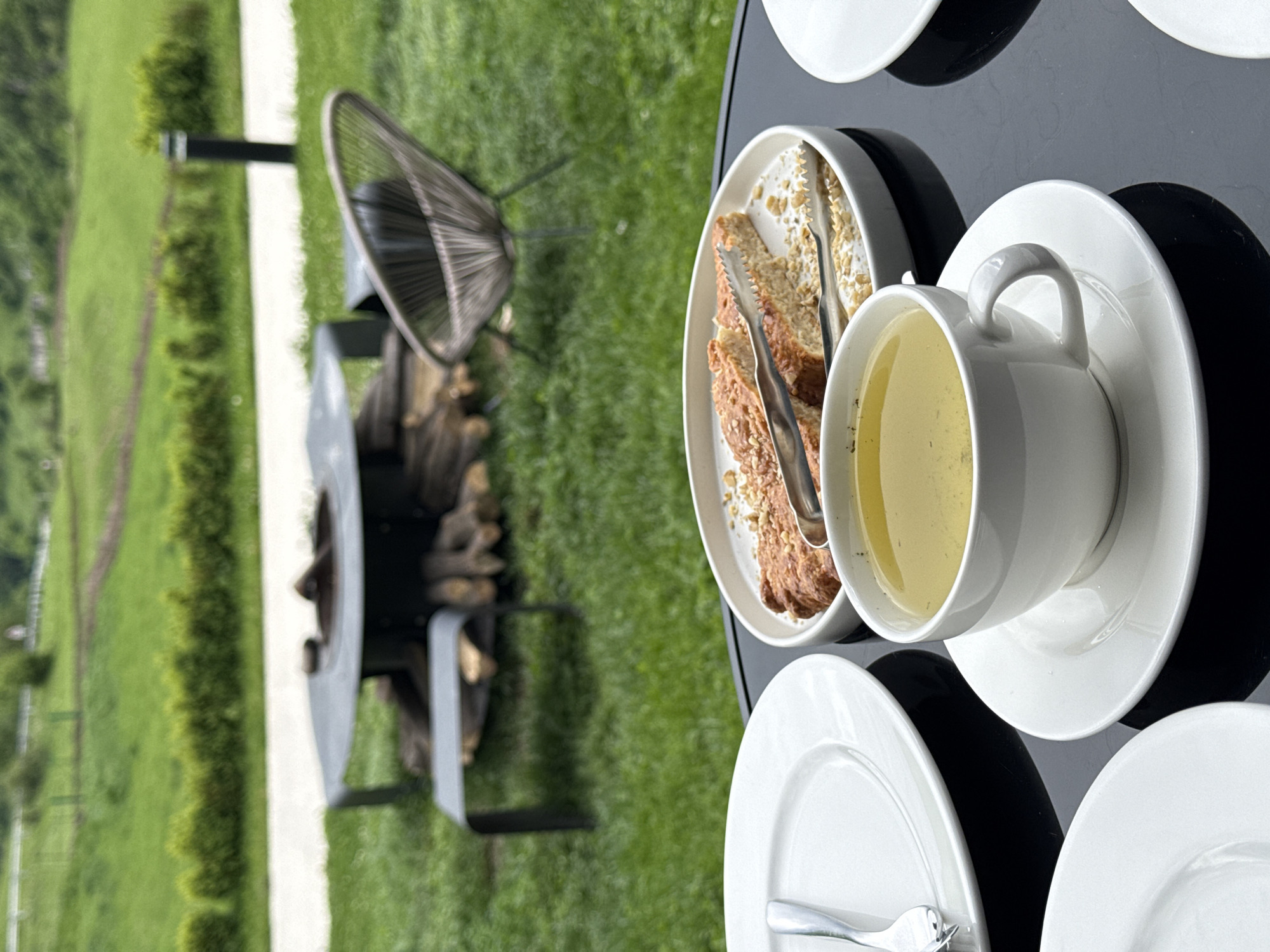 A close-up, rotated image depicts a black table on a grassy area, likely in the vicinity of Chkalov, Armenia, given the mountainous backdrop.
On the table, there are several white plates and a teacup filled with a yellow liquid (presumably tea). A plate holding slices of cake is also present, accompanied by a pair of serving tongs.
In the background, a fire pit with wood stacked inside stands next to a chair with a woven seat. A bright green lawn stretches out, and a white stone pathway edges the area. The image seems to capture a moment of outdoor dining or a break with a view of the surrounding landscape.
