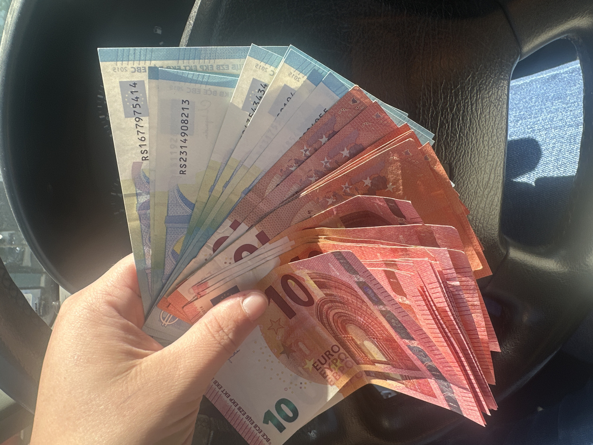 A hand holds a fan of Euro banknotes, comprised of ten-Euro, twenty-Euro, and five-Euro denominations, against the backdrop of what appears to be the interior of a vehicle, with a steering wheel visible. The denominations are mixed and the arrangement suggests a collection of various values of European currency. There is no explicit information to place this image in the city of Rakovski, Bulgaria.