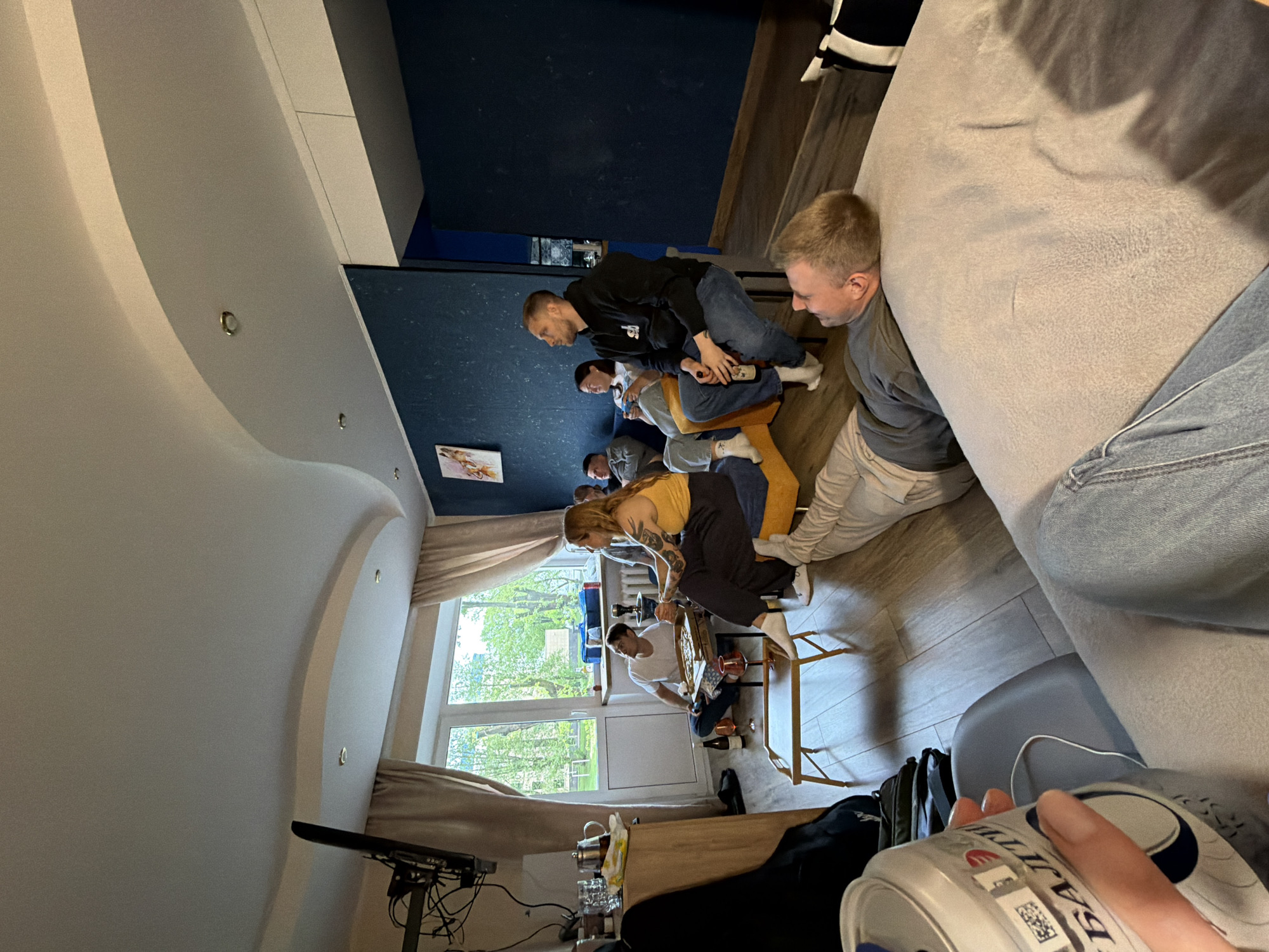 A low-angle, interior shot shows a group of people gathered in a room. Six individuals are seated in a circle. A person dressed in a yellow shirt is seated, facing the camera, while another person across from them is working on an unknown object. A man in a black sweatshirt sits behind them. In the foreground, a hand holds a beverage can. The room is illuminated by natural light coming through a large window, which reveals a leafy, outdoor scene. A television and other items are visible on a shelf near the window. The walls are painted a dark color, and a piece of artwork is hanging. A sofa is visible, occupying space in the foreground of the frame.