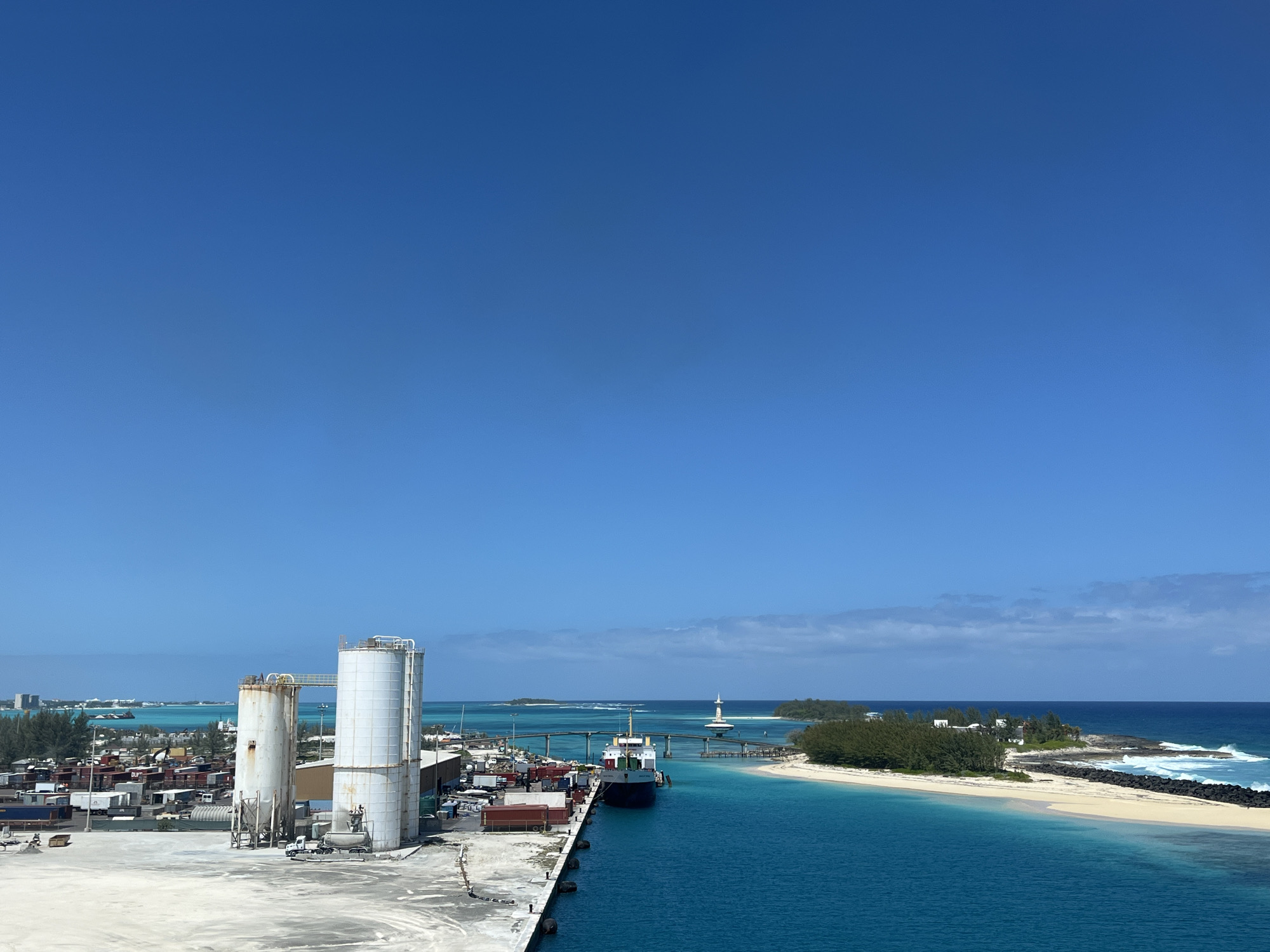 The image shows an elevated view of a port area. In the foreground are two large white silos next to a concrete pier lined with shipping containers and other cargo. A cargo ship is docked at the pier. A narrow channel of turquoise water leads out to the ocean. A small island with a white sandy beach, vegetation, and a breakwater is on the right side of the channel. A lighthouse stands on a small island in the channel. Beyond that is the ocean under a clear blue sky. In the far distance, the skyline of a city is visible on the horizon. The location is in the city of Unknown, Unknown.