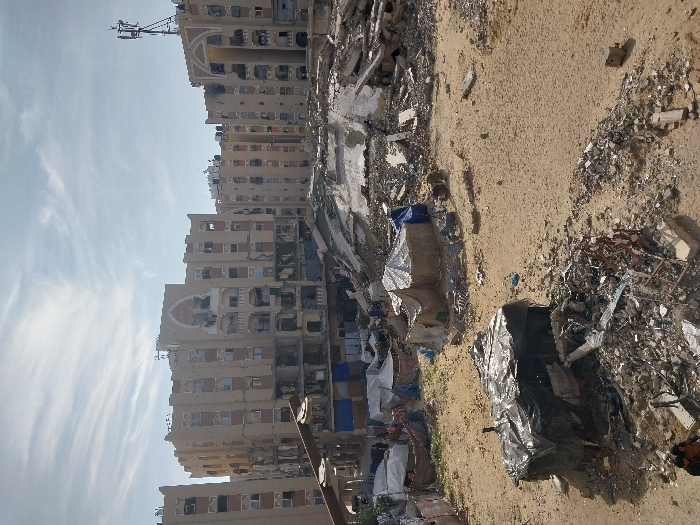 This image depicts a wide-angle view of a heavily damaged urban environment in Khan Yunis, Palestinian Territory, under a partly cloudy daytime sky.
In the background, a row of multi-story, light-colored apartment buildings stands. These buildings exhibit extensive structural damage, including numerous shattered windows, missing wall sections exposing interiors, large impact craters, and blackened areas consistent with fires or explosions. Debris hangs from various floors, and some buildings feature decorative arched elements near their rooftops. A communications tower, possibly a cellular antenna array, is mounted on the roof of a building on the far right.
The midground is characterized by a vast expanse of destroyed infrastructure. This area is filled with concrete rubble, twisted metal rebar, collapsed building fragments, and general debris, indicating significant demolition or destruction. Amidst this devastation, particularly towards the left, a cluster of makeshift shelters and tents is visible. These temporary structures are constructed from various materials, including tarpaulins (some blue, some dark), fabric, and scavenged sheets, appearing rudimentary and erected directly on the sandy ground or amidst smaller debris piles.
The foreground consists of barren, sandy soil interspersed with scattered debris. On the lower left, a dark, box-like structure covered with black plastic sheeting or tarpaulin stands amidst a concentrated pile of fragmented concrete, twisted metal, and assorted refuse. In the immediate lower-left corner, two individuals are visible, seated or crouching near this debris pile, their attire indistinct due to distance and resolution. The overall ground surface appears dusty and uneven.