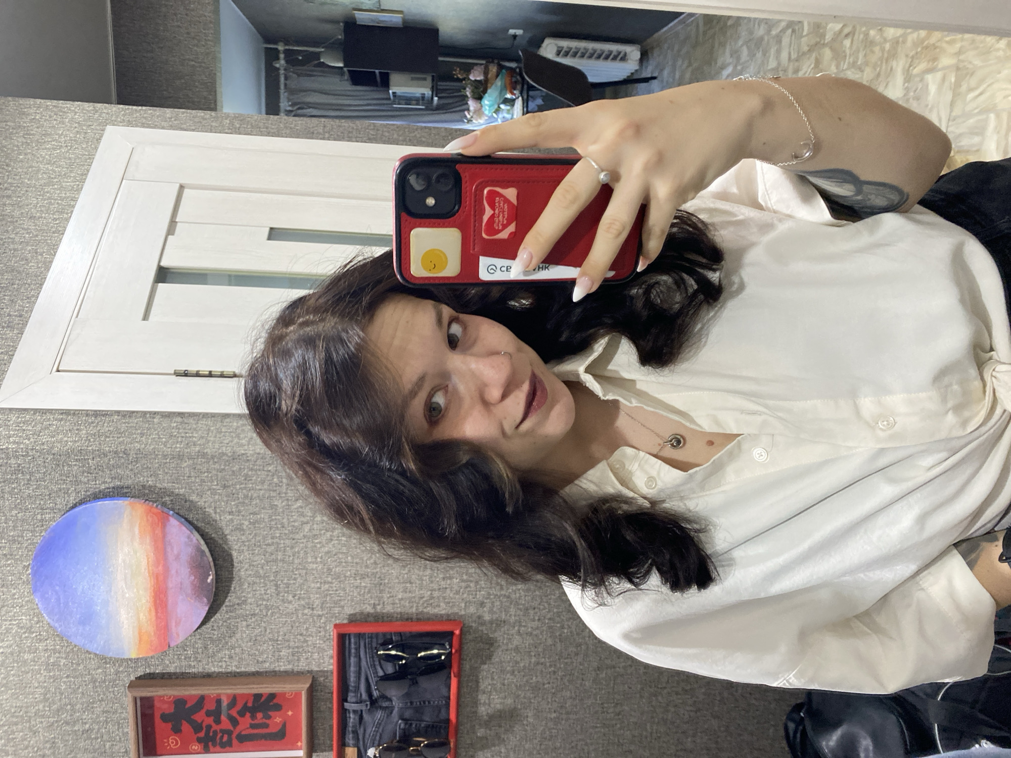 A woman with dark, wavy hair and light skin is taking a selfie in a room. She is wearing a white button-up shirt and dark pants. She has a ring on her left ring finger and a bracelet on her left wrist. She is holding a red phone in her right hand. On the wall behind her are two framed items, one with red lettering and another with a depiction of a sunset. A white door is visible to her right. The room appears to be a domestic setting, possibly an apartment in Chaikovsky, Russia, given the context.