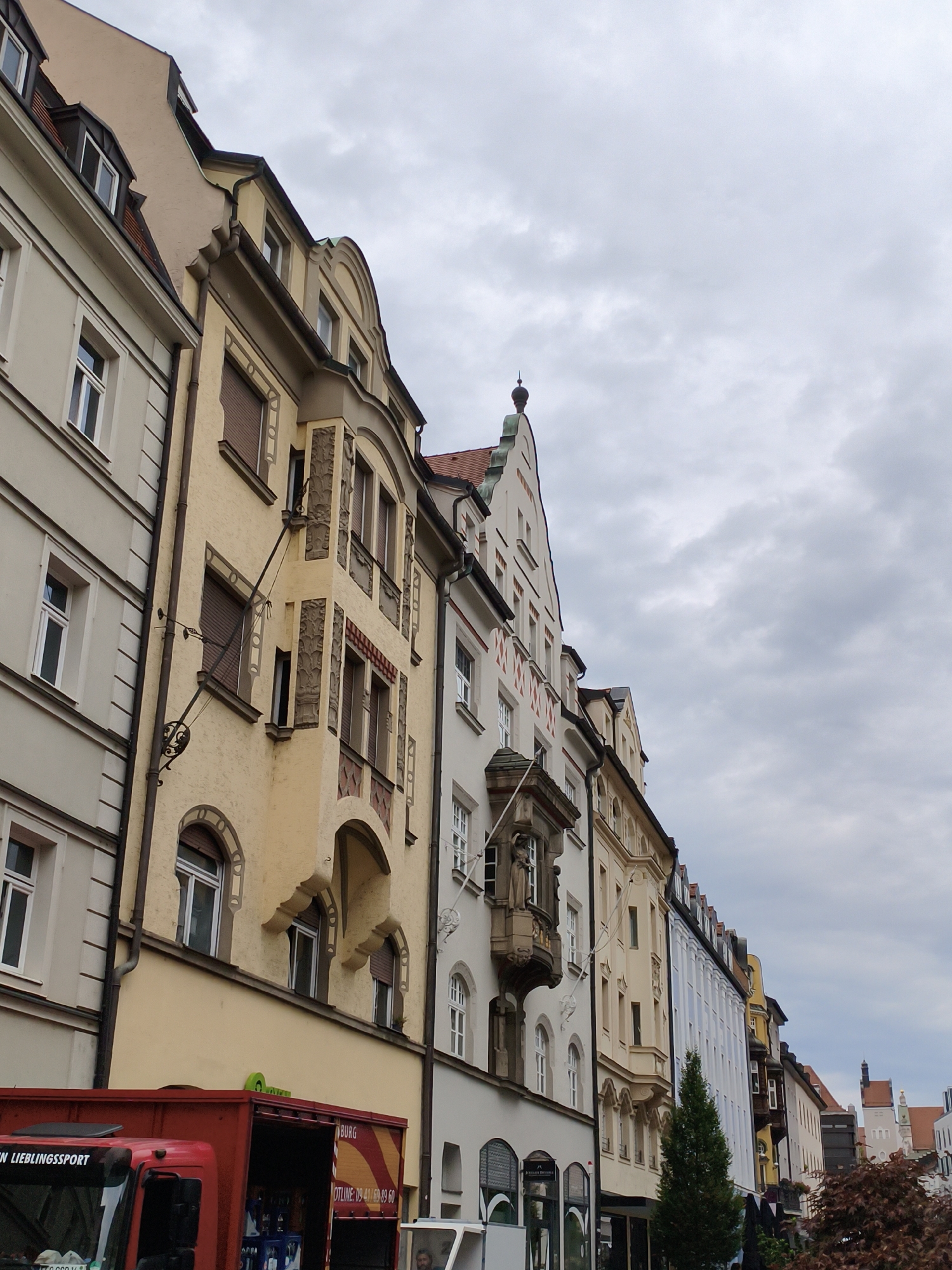 The image shows a street scene in Regensburg, Germany, looking up at a row of historic buildings. The architecture is diverse, with several styles represented, including some with ornate facades and mansard roofs. A red truck is visible in the foreground, partially obscuring some of the lower floors of the buildings. The sky is overcast, with dramatic clouds. The scene appears to be a typical urban environment, with the focus on the architecture and the muted daylight.