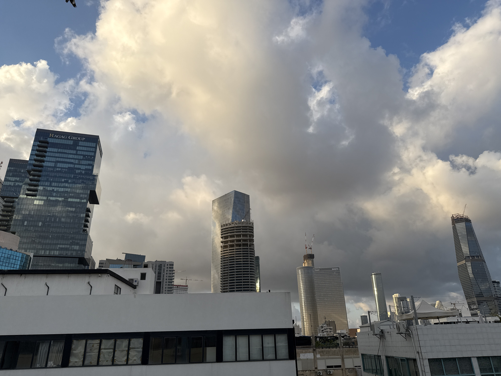 The image presents a modern cityscape in Tel-Aviv, Israel, dominated by towering skyscrapers under a dramatic, cloudy sky. The scene is viewed from a slightly elevated position, looking up over a flat, white-roofed building in the foreground, which features dark-framed windows.
Several prominent buildings shape the skyline. On the far left, a dark, multi-story glass building displays the text "HAGAG GROUP" clearly visible at its top, identifying a key player in the Israeli real estate market. In the center, a tall, slender building is under construction, its rounded, concrete skeleton exposed against the sky, with another highly reflective skyscraper shimmering behind it. Further to the right, an extremely tall, sleek building, also appearing to be under construction, ascends skyward, topped with cranes. Numerous other construction cranes are scattered across the urban landscape, signifying ongoing development.
The sky is a focal point, characterized by a mix of bright blue patches and large, impressive white and grey clouds. The clouds exhibit warm, yellowish hues in places, suggesting the soft, low light of either late afternoon or early morning. A small dark bird is visible flying high in the upper left corner. The overall impression is that of a vibrant and growing metropolis.