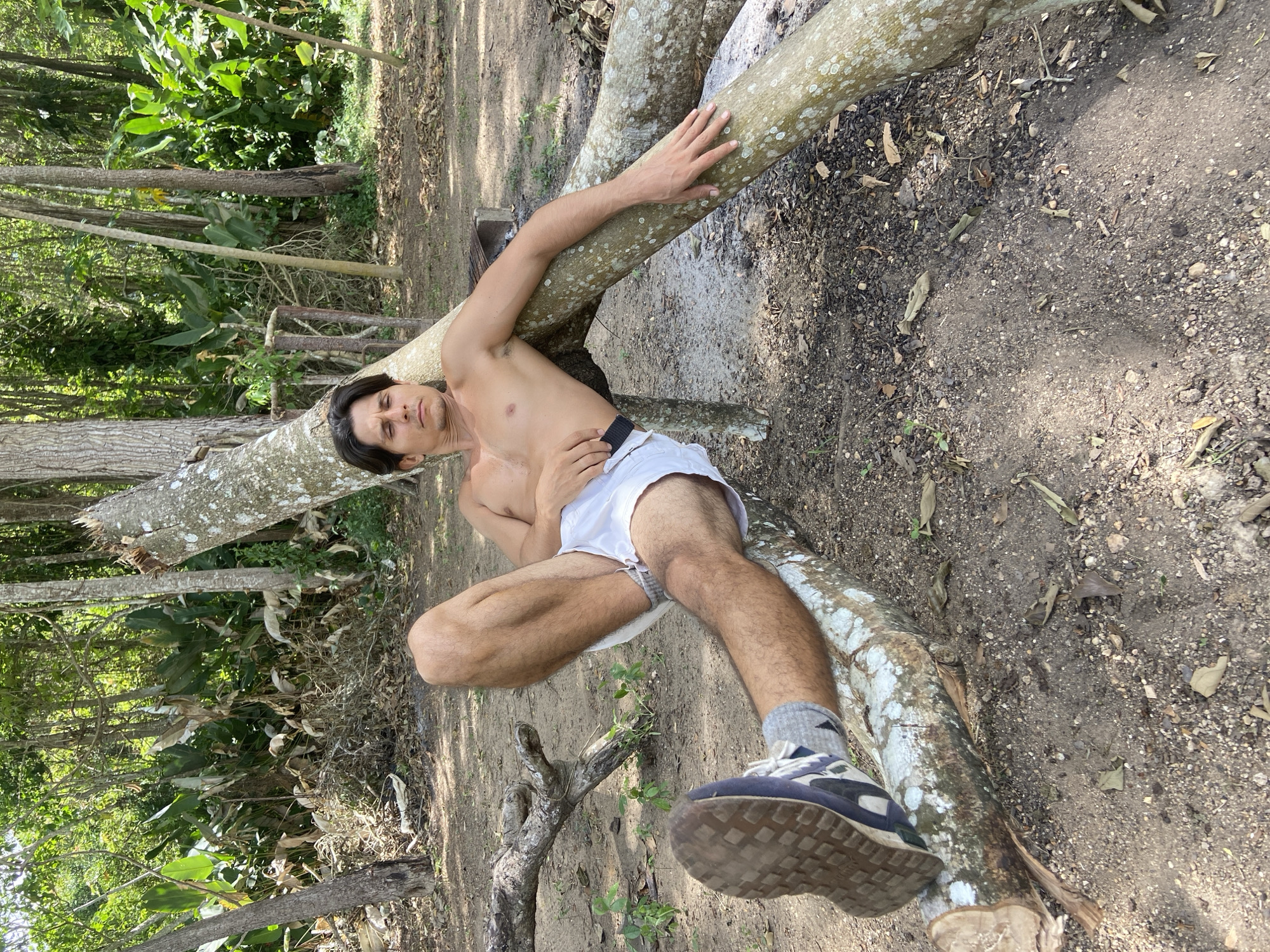 A shirtless man with dark hair and a light complexion is reclining on fallen tree branches in a wooded area. He is wearing white shorts and grey socks with blue and black athletic shoes. His left leg is extended, and his right arm is resting along a branch. The ground is covered in dirt, small stones, and fallen leaves. In the background, there are trees and dense vegetation. The lighting suggests it is daytime with sunlight filtering through the trees. The scene is outdoors in a natural environment, possibly a park or forest area within Los Velásquez, Venezuela.