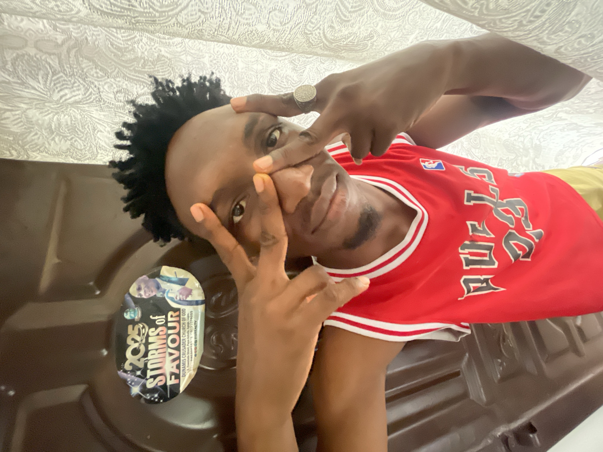 The image is a selfie of a young man with dark skin, wearing a red basketball jersey with white stripes. He is making a gesture with his hands near his face. The background is a patterned white fabric, possibly a curtain or sheet. A sticker with the words "2025 STORMS of FAVOUR DUNAMIS CRUSADER CHURCH OF GOD" is visible on a dark surface, likely a table or door. The sticker provides information about the Dunamis Crusader Church of God and an address in Lagos, Nigeria. There is no mention of the city of Lokoja in Nigeria.