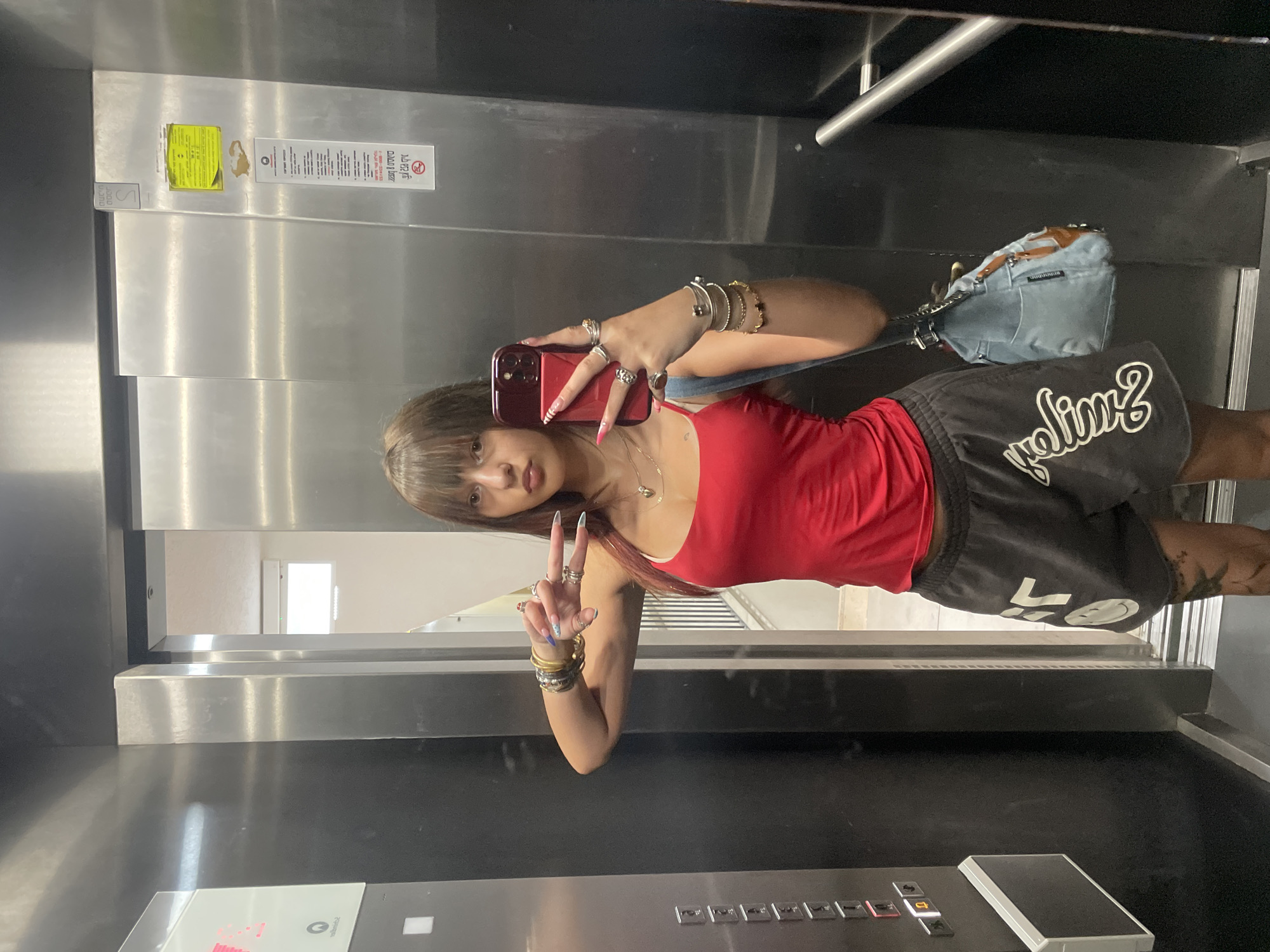 A young woman with bangs and dark brown hair is standing in an elevator, taking a mirror selfie with a smartphone. She is wearing a red tank top, grey shorts with white lettering and graphics, and numerous bracelets and rings on her arms and hands. She is holding up two fingers in a peace sign with her left hand. A light blue shoulder bag with brown leather accents is slung over her right shoulder. The elevator has stainless steel walls and black paneling with illuminated buttons on the left side. A yellow sticker with text and a red symbol is affixed to the elevator wall on the right. The elevator doors are partially open, revealing a glimpse of a building interior with a staircase and windows. The scene is indoors, likely in an apartment building or commercial space in Raanana, Israel, given the context provided.