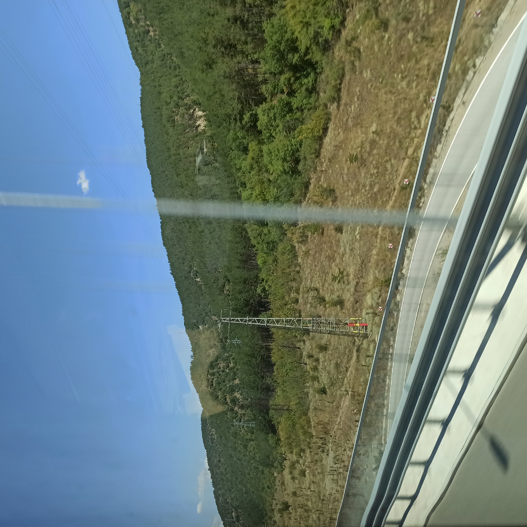 The image depicts a scenic view of a hillside covered in dense green trees, with a clear blue sky above. A transmission tower stands prominently on the hillside, with power lines stretching across the sky. A road, with a guardrail, curves through the landscape in the foreground. The overall impression is one of a natural environment with man-made structures integrated into the landscape. The scene is likely captured from a moving vehicle, given the slight motion blur evident in the foreground.