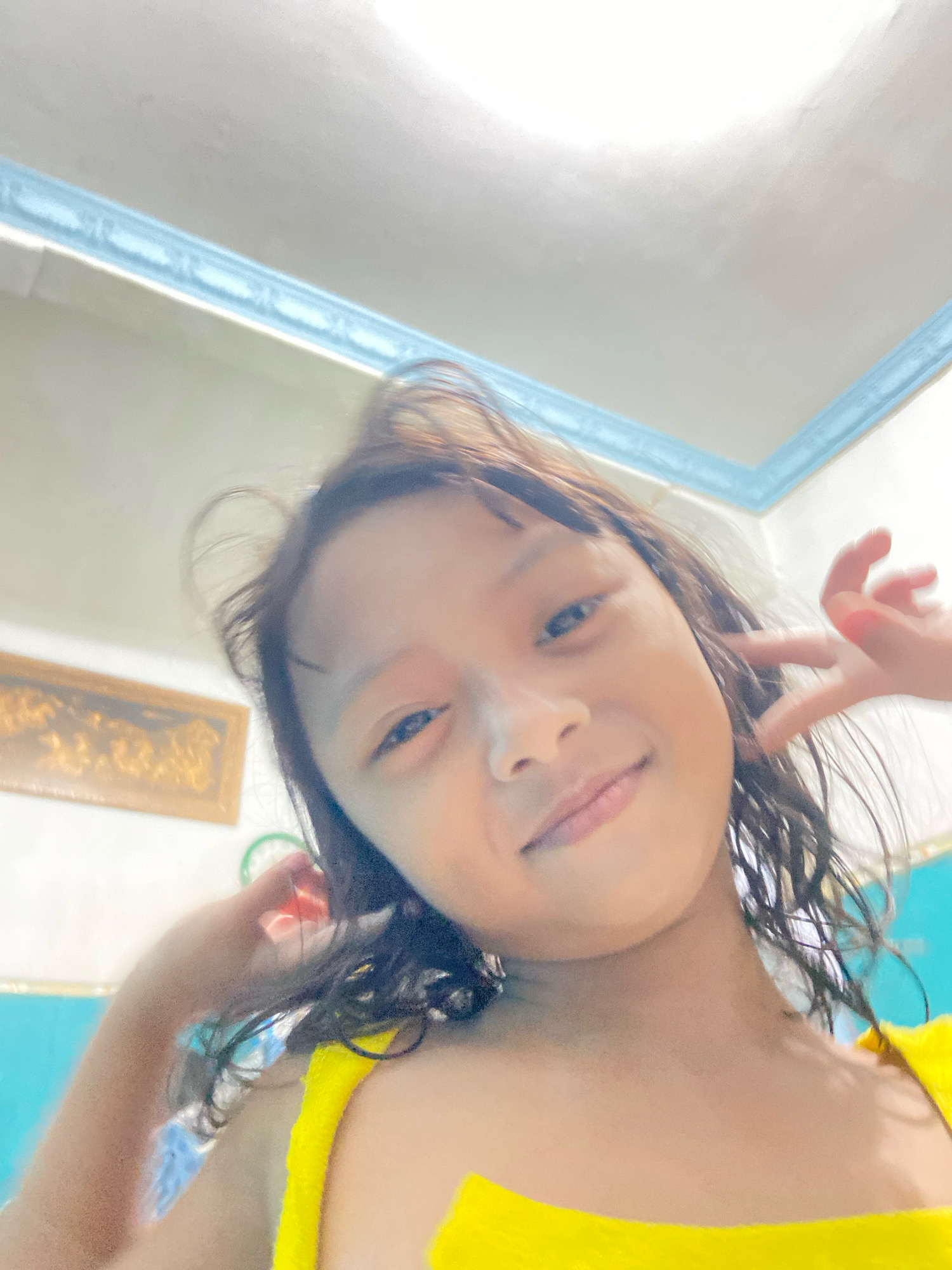 This is a close-up selfie of a young girl in Gelam, Indonesia. She has dark, wavy hair and is smiling at the camera with her left hand near her face. She is wearing a bright yellow top. The background shows a white ceiling with a light blue decorative trim. To the left of the girl, a framed picture with a golden frame is visible on the wall. The overall lighting suggests it is daytime, possibly indoors with bright overhead lighting. The girl appears happy and playful, with a relaxed and engaging expression. No visible text is present in the image.