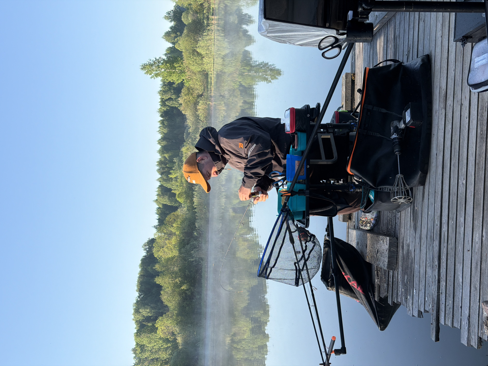 A man wearing a brown baseball cap and a black jacket is fishing from a wooden dock. He is seated on a blue chair equipped with various fishing accessories, including fishing rods and a net. The dock extends into a body of water with a forested shoreline on the opposite bank. The water is calm, reflecting the trees and sky. The sky is clear and blue. The location is Belaya Gora, Russia.
