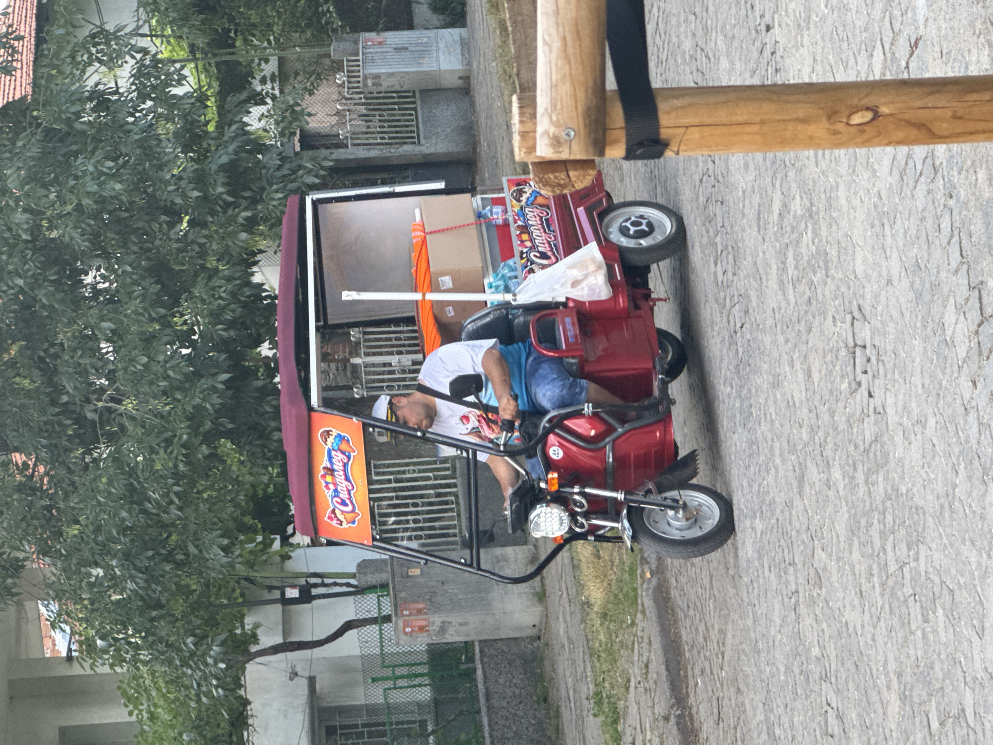 A man is driving a red, three-wheeled motorized cart down a cobblestone street in Plovdiv, Bulgaria. The cart has a canopy overhead and is equipped with a serving area. The side of the cart features an advertisement for "Cragoregg" ice cream, depicted with cartoon illustrations of ice cream cones and a sun. The man is wearing a white t-shirt with an ice cream design, blue shorts, and a white cap. He is holding onto the handlebars, steering the vehicle. The cart is carrying various items, including boxes, bottles of water, and a white plastic bag. The background shows buildings, trees, and a metal fence, typical of a city environment. The overall scene captures a moment of daily life, likely involving the sale of ice cream from the cart.