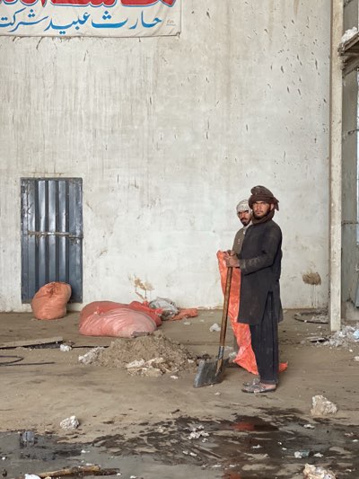Two men are standing in what appears to be a large, unfinished or dilapidated building. The man in the foreground is holding a shovel and is wearing dark traditional clothing and a turban. A second man stands slightly behind him, also wearing traditional clothing and a head covering. In the background there are several large bags, a barred window, and debris scattered across the floor. A sign with Arabic script is visible near the top of the image. The scene is set in Kandahar, Afghanistan.