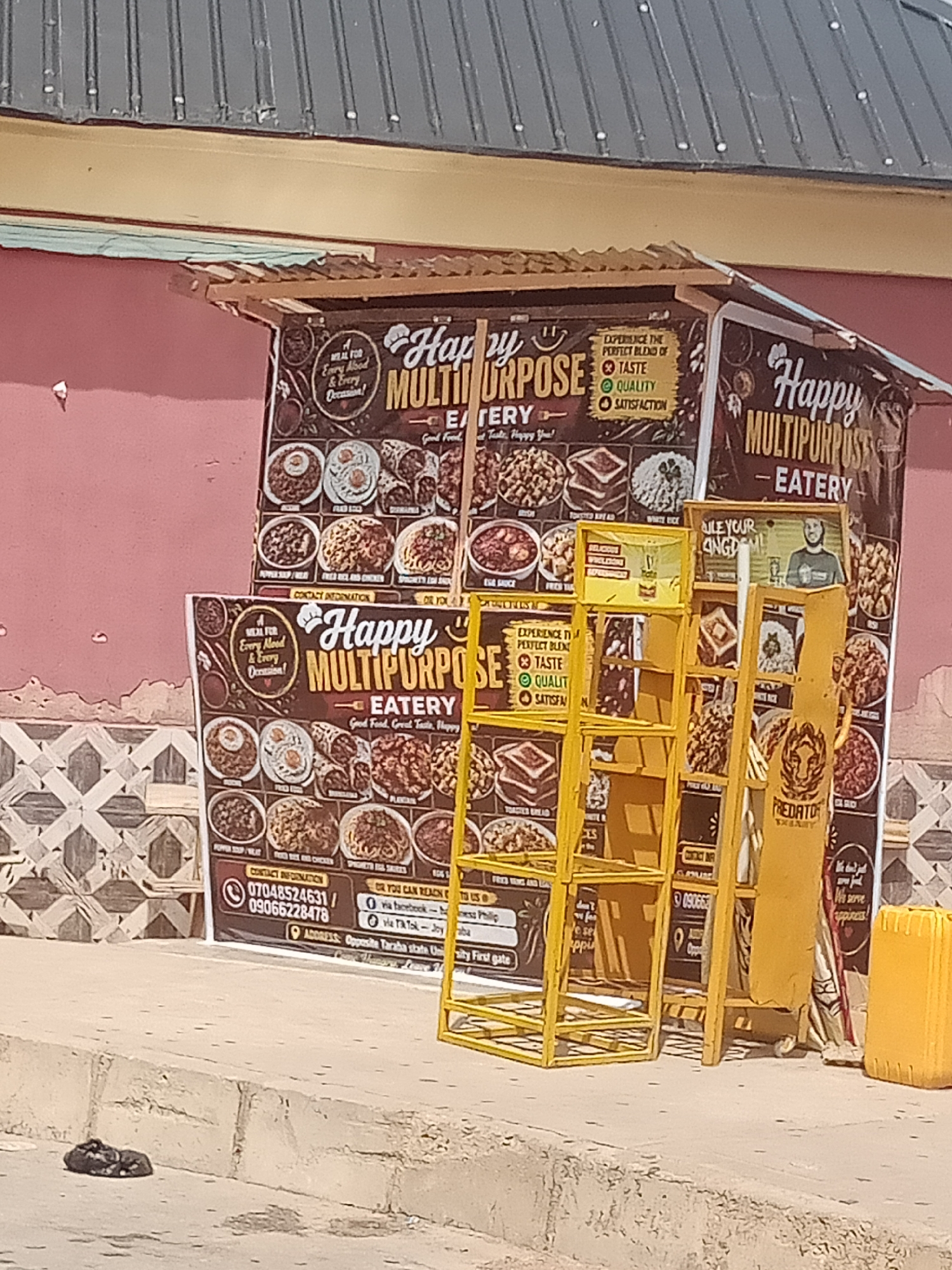 This media file depicts an outdoor food stall in Jalingo, Nigeria. The stall, named "Happy Multipurpose Eatery," is constructed with a corrugated metal roof and a pink-painted wall as its backdrop. It features prominent signage displaying a variety of food items and promotional text.
The primary focus is the food stall itself, adorned with colorful menus and a yellow display rack holding what appear to be food samples or advertisements. The menus showcase images of dishes like pepper soup, fried rice and chicken, spaghetti, and toasted bread. Text on the signage emphasizes "Taste, Quality, Satisfaction" and offers contact information, including phone numbers and social media handles.
Adjacent to the stall, a yellow plastic container and a rolled-up mat are visible. The ground in front of the stall is a concrete sidewalk, and a small, dark object, possibly a discarded piece of clothing or trash, lies on the ground to the left. The scene suggests an active commercial setting, likely during daylight hours given the clear illumination. There are no people clearly visible in the image, but the presence of the food stall and its promotional materials indicates a place where food is being sold and customers are expected. The overall impression is of a modest, local business operating on a street.