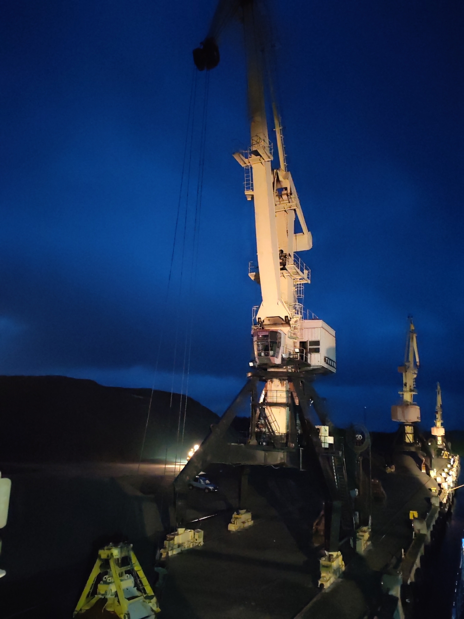 The image shows several large port cranes against a dark blue evening sky. The location is presumably in Shalsky, Russia, given the prompt. One primary crane is centrally positioned, its white structure illuminated against the night sky. Supporting cables descend from its arm. Other cranes are visible in the background, along a loading dock or pier. The area below the cranes is filled with dark materials, possibly coal or ore, with concrete supports visible. The backdrop is a dark landmass or mountain range. Some ambient lights illuminate the scene, and a vehicle is partially visible.