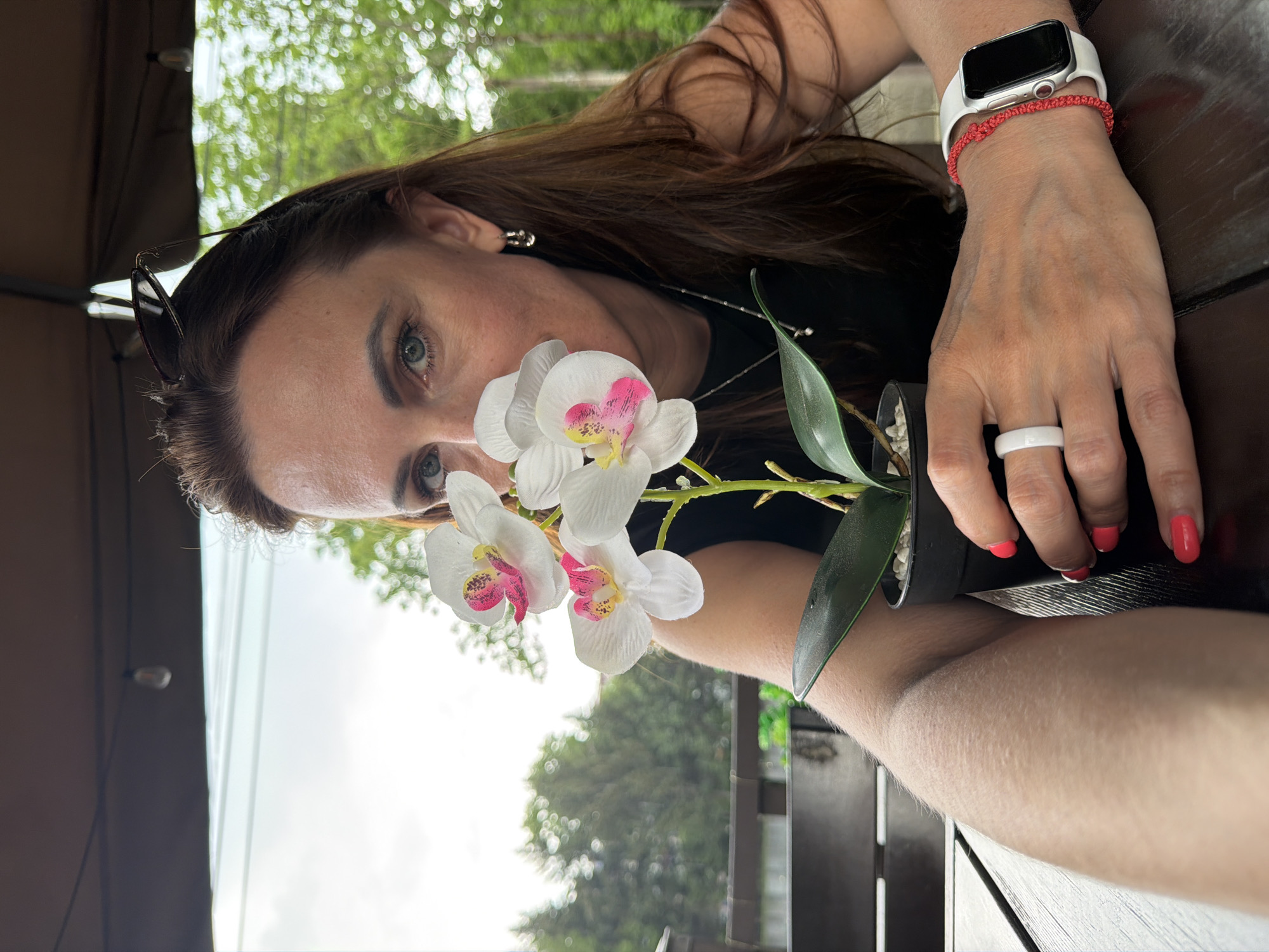 A woman is captured from the chest up, holding a potted orchid in her left hand and bringing it close to her face, partially obscuring it. She has long, dark brown hair and blue eyes, and is wearing a black top, silver hoop earrings, a white ring on her left ring finger, and a red bracelet with a white Apple Watch on her left wrist. Her fingernails are painted bright pink. The background suggests an outdoor setting, possibly a cafe or restaurant patio, with a wooden table in the foreground and blurred trees and a cloudy sky visible behind her. The scene is set during daytime. The location in Birobidzhan, Russia, is not discernible from the image.