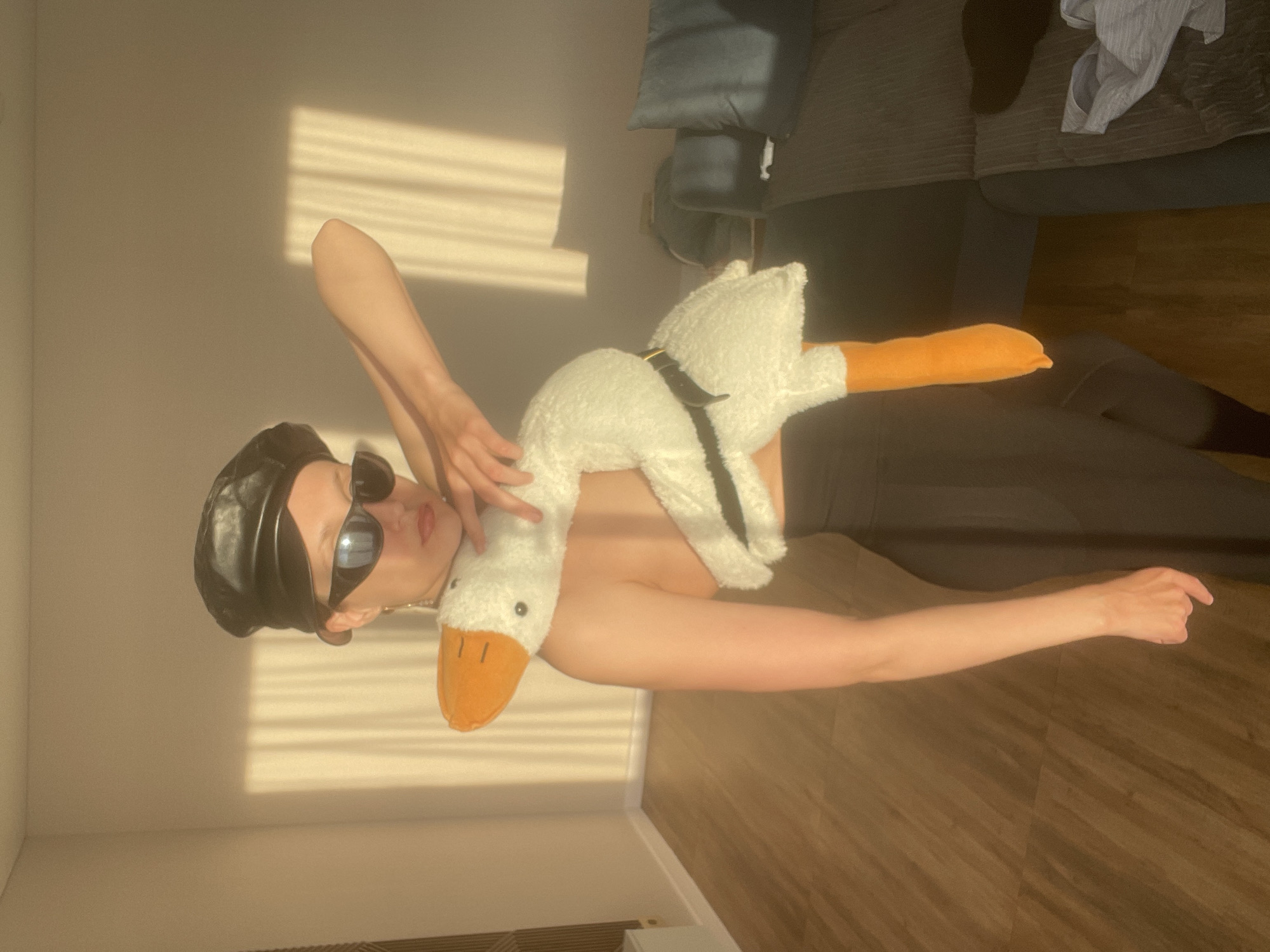 An indoor, three-quarter shot captures a person posing with a large white plush goose toy. The image is rotated 90 degrees clockwise. The subject is light-skinned, wearing a black leather beret and dark sunglasses that obscure their eyes. Their lips are slightly parted. They are dressed in a black top or dress, possibly strapless, and black trousers or leggings. One arm is raised, holding the goose's head, while the other arm is extended towards the lower right of the frame.
The plush goose has a fluffy white body, an orange beak, black eyes, and orange feet. A black strap or harness detail is visible around its back.
The setting is a room with light-colored walls and a dark wood laminate floor. Bright sunlight streams through a window, casting prominent horizontal shadow lines from window blinds across the wall and floor, indicating it is daytime. In the upper right background, a dark sofa or armchair with cushions is visible, on which a light grey piece of clothing rests. The overall scene suggests a casual, stylized pose in a sunlit interior in Podshivalovo, Russia. No visible text is present.