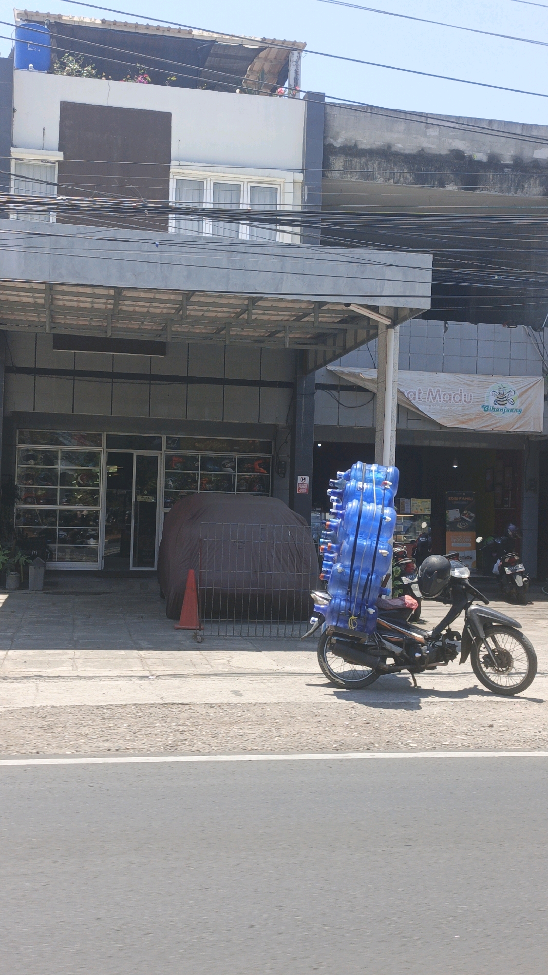 The image shows a street scene in Pangandaran, Indonesia, on a sunny day. In the foreground, a black motorcycle is parked on the side of the road, loaded with a tall stack of blue plastic water jugs. The motorcycle has a black helmet on its handlebars. Behind the motorcycle, a brown tarp covers a car parked in front of a shop. The shop has a large storefront with many windows displaying various items. A red traffic cone is placed on the sidewalk near the covered car. Above the shop, an awning provides shade. Further up, the upper floors of buildings are visible, with windows and some greenery on a rooftop. Electrical wires are strung across the scene at various levels. In the background, other motorcycles and parts of buildings can be seen, along with a banner that reads "Cihanjuang" and "Jual Madu" (Honey for Sale). The overall environment suggests a commercial area with ongoing activity.