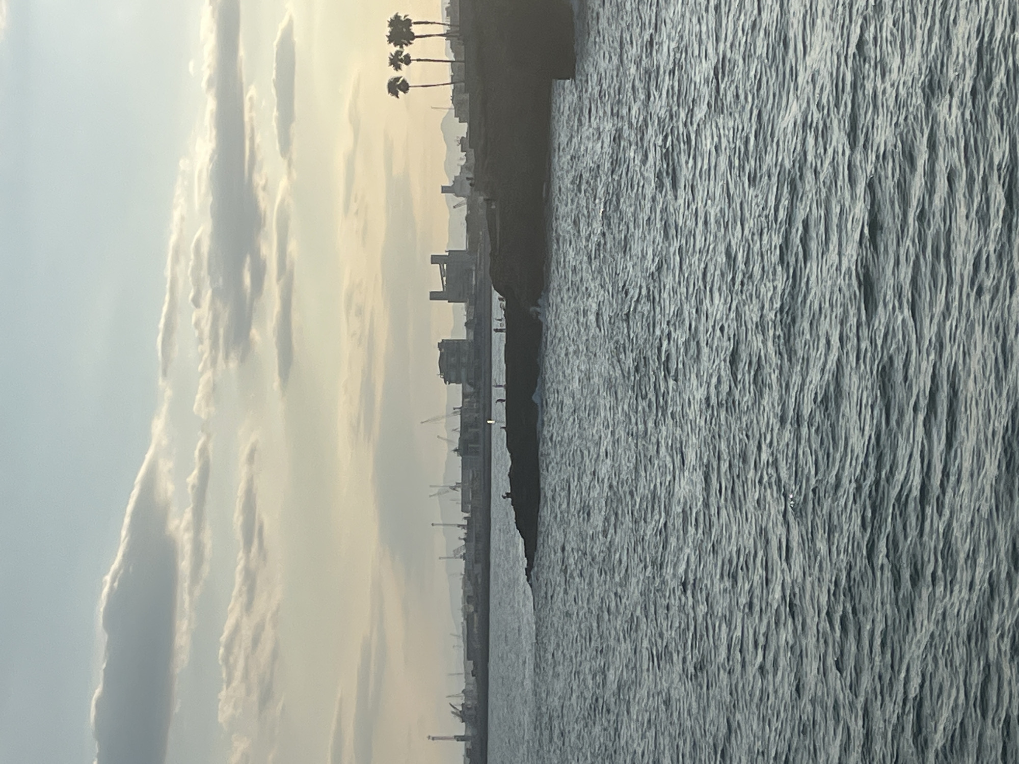 The image shows a coastal scene in El Miracle, Spain. The foreground is dominated by the rippling surface of the sea. In the mid-ground, a rocky outcrop extends into the water, with several silhouetted figures standing on it. Beyond the outcrop, a harbor skyline is visible, characterized by numerous cranes and industrial structures. A few palm trees stand on the right side of the frame, silhouetted against the hazy sky. The sky itself is a soft gradient of pale blue and yellow, with scattered clouds, suggesting either dawn or dusk.