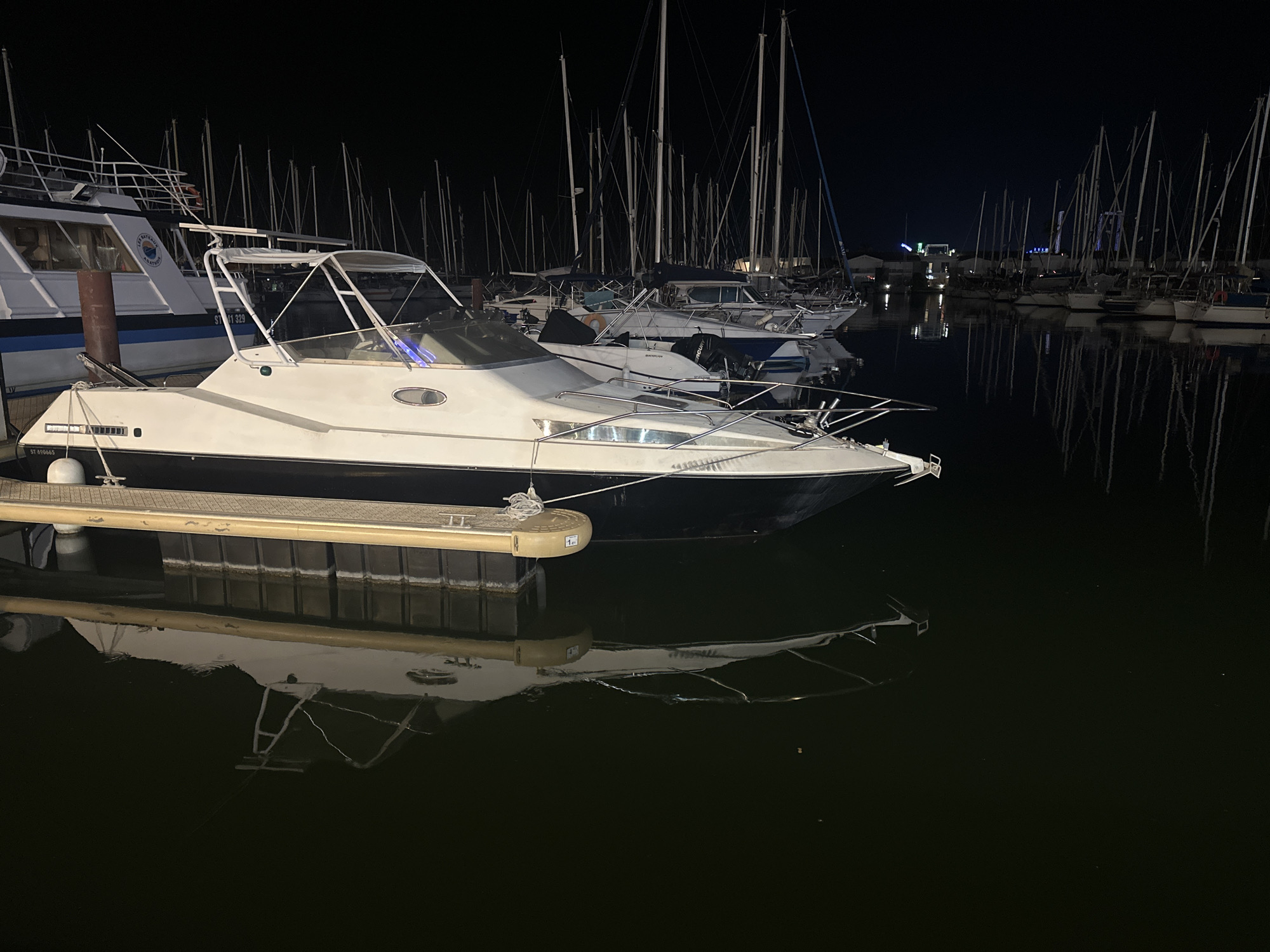 At night, several boats are docked in a marina in Agde, France. The foreground features a white motorboat with a black hull, its reflection visible in the dark water. Behind it, numerous sailboats with tall masts are moored. The marina is illuminated by artificial lights, creating reflections on the water's surface. In the background, distant lights suggest the presence of buildings or structures on land. The overall scene depicts a tranquil night at a French marina.