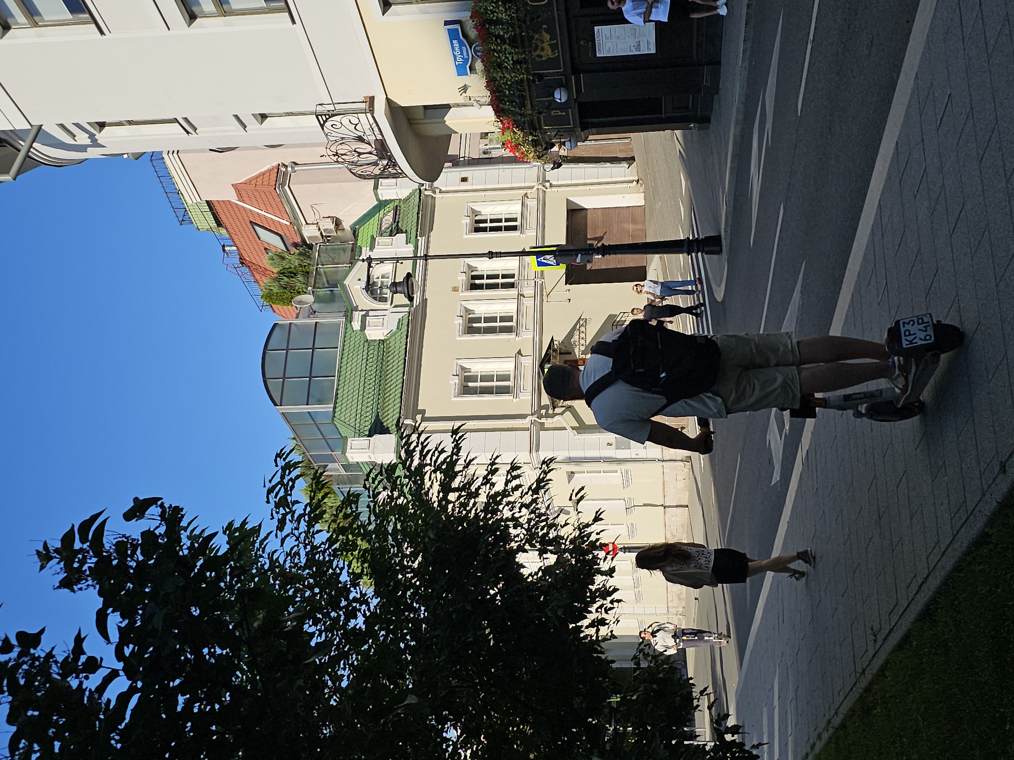The image depicts a sunny day on a city street, likely in Moscow, Russia, given the Cyrillic street sign reading "Трубная улица" (Trubnaya Street). In the foreground, a person rides an electric scooter, viewed from behind. The rider wears a backpack and light-colored shorts. Further down the sidewalk, a woman in a black skirt and a light-colored top walks away from the camera. Another person on an electric scooter is visible in the middle ground on the left side of the street. The street itself is paved and lined with buildings.
The buildings have varied architectural styles. On the left, a light-colored building with multiple windows and a green roof is prominent. On the right, a building with a curved facade and a black awning over an establishment labeled with Cyrillic text, possibly a pub or restaurant, is visible. Above this, an ornate black metal balcony adds to the architectural detail. The sky is clear and blue. The overall impression is one of a lively urban environment on a pleasant day.