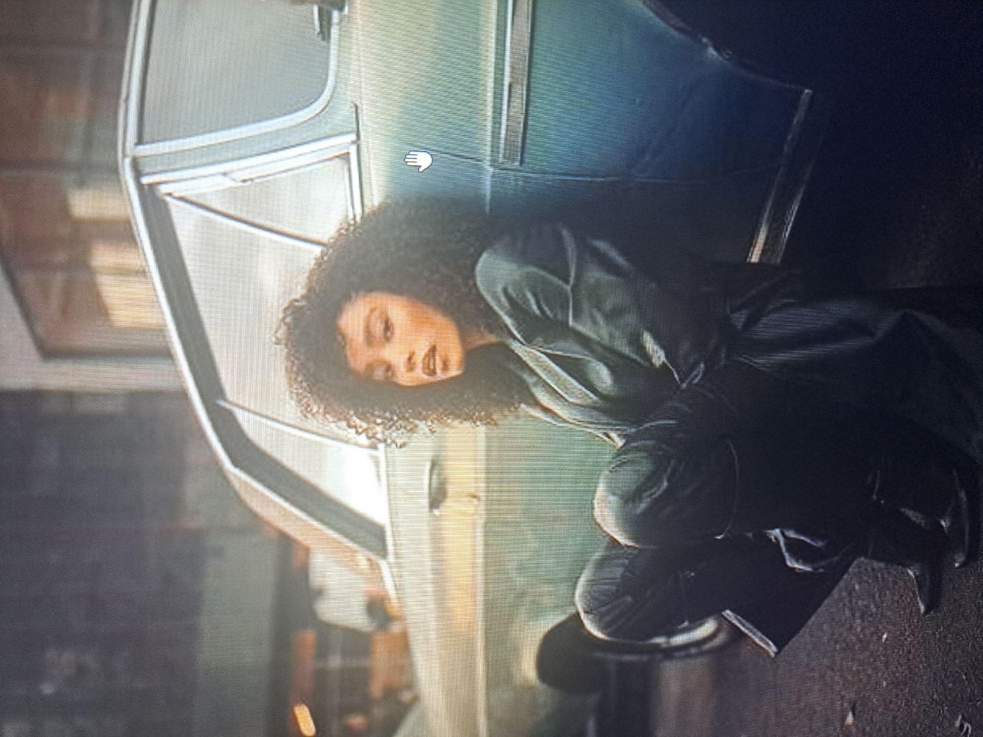 A woman with curly hair is crouching near a light blue vehicle, possibly a bus or van. The setting appears to be urban, with building facades visible in the background, suggestive of a city environment. The image does not contain enough information to determine whether the scene is in Yerevan, Armenia.