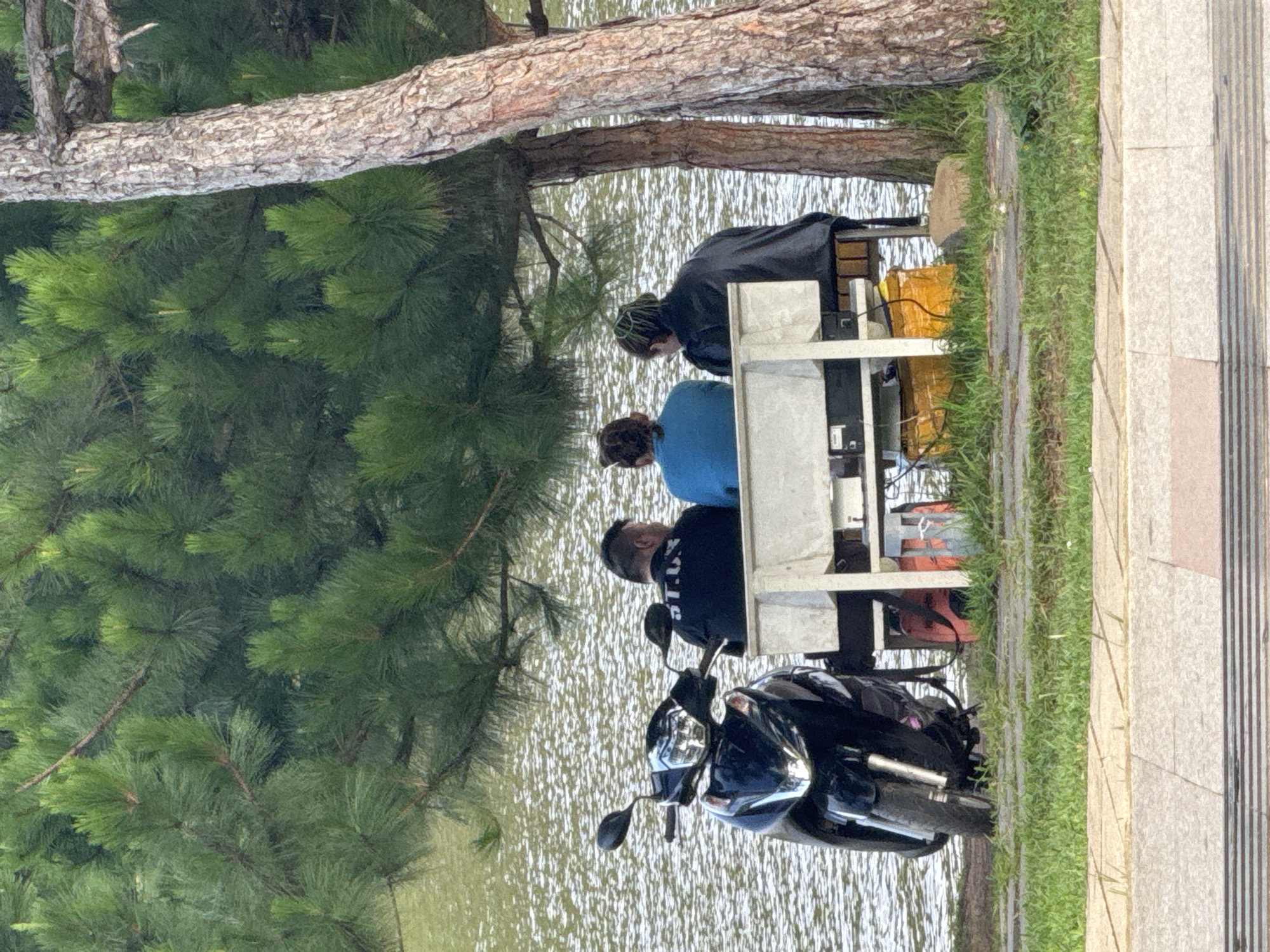 Three individuals are seated on a white bench next to a body of water, likely Xuan Huong Lake in Da Lat, Vietnam, under the shade of pine trees. A black scooter is parked to the left of the bench. The individuals, facing away from the camera, are dressed casually. The person on the left wears a black long-sleeved shirt with white lettering, the middle person wears a bright blue long-sleeved shirt, and the person on the right wears a dark jacket. Beneath the bench, several items are visible, including orange and yellow bags. The water has a rippled texture, suggesting a gentle breeze. The surrounding area is covered in grass and paved pathways. The lighting indicates it is daytime, possibly late afternoon given the dappled sunlight filtering through the trees.