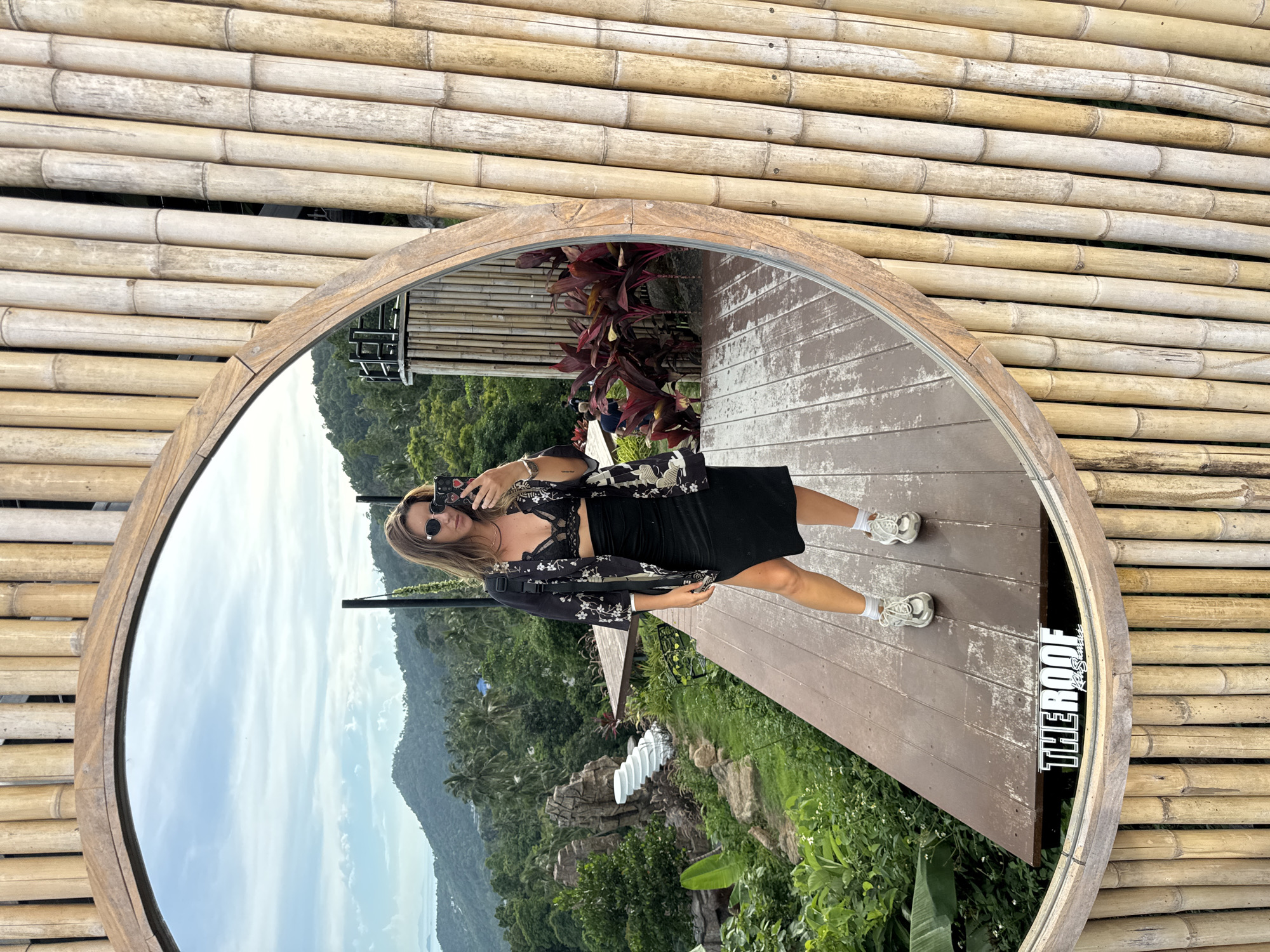 The image captures a woman taking a mirror selfie in an outdoor setting, likely a balcony or deck, surrounded by bamboo stalks. The woman is wearing sunglasses, a black lace bralette, a black skirt with a slit, a black patterned kimono-style jacket, white socks, and white sneakers. She is holding a smartphone in her right hand. In the reflection of the mirror, a lush, mountainous landscape with palm trees and a clear sky is visible. The foreground of the reflection shows a wooden deck with white painted accents. A sign that reads "THERROOF" is visible on the deck. The overall atmosphere suggests a tropical or exotic vacation destination.