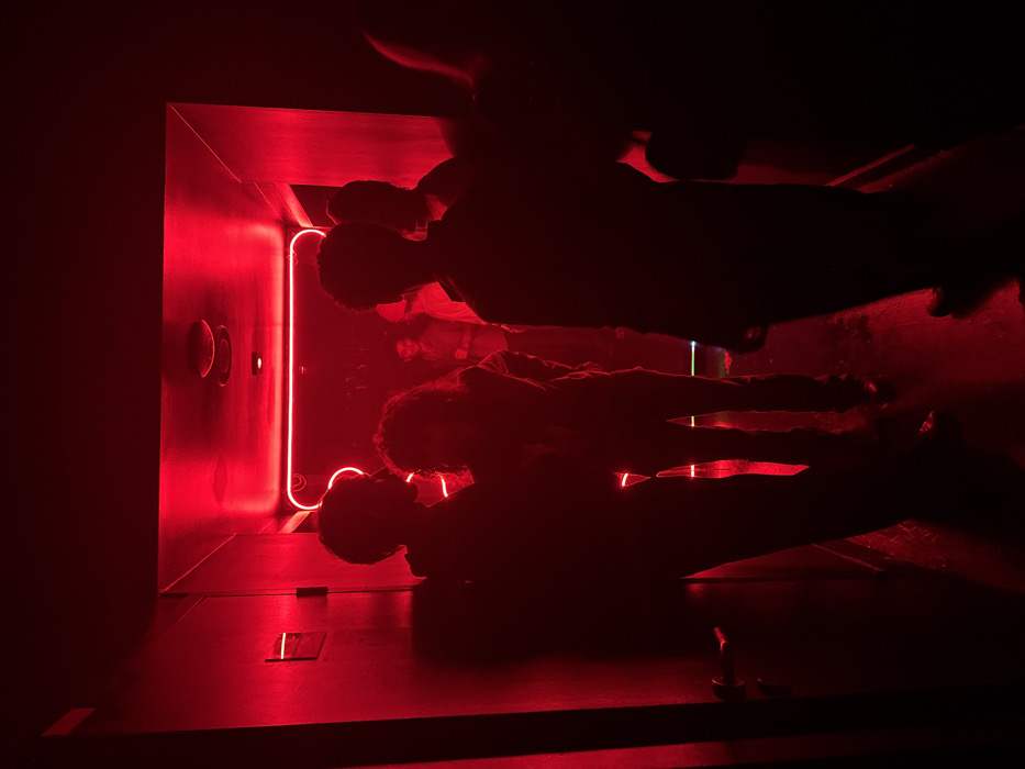 A group of people is illuminated in a dark space with intense red lighting. The subjects appear to be conversing within a compact, enclosed area, possibly a small room or chamber. A vibrant red neon strip outlines a portion of the space, casting harsh shadows and highlighting the silhouettes of the individuals. The focus is on the interplay of light and shadow, creating a dramatic and intimate scene.