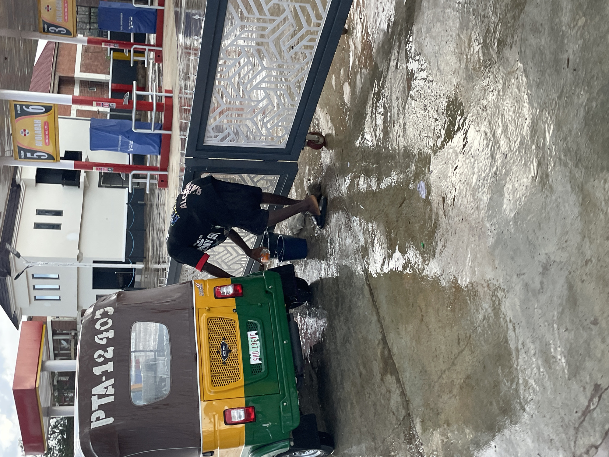 A person wearing a black t-shirt and dark shorts is washing a yellow and green auto-rickshaw at a gas station in Barakin Lerawi, Nigeria. The auto-rickshaw has "PTA-12403" painted on its canopy and a license plate reading "SHD-196YC". The person is holding a sponge and appears to be washing the vehicle. A blue bucket is on the ground next to the auto-rickshaw. The ground is wet and reflective, suggesting recent rain or washing. In the background, there are gas pumps with signage indicating "PUMP 5" and "PUMP 6" from AI Olabah. A white building with a red roof is also visible behind the gas station. A decorative metal gate is positioned to the right of the auto-rickshaw.