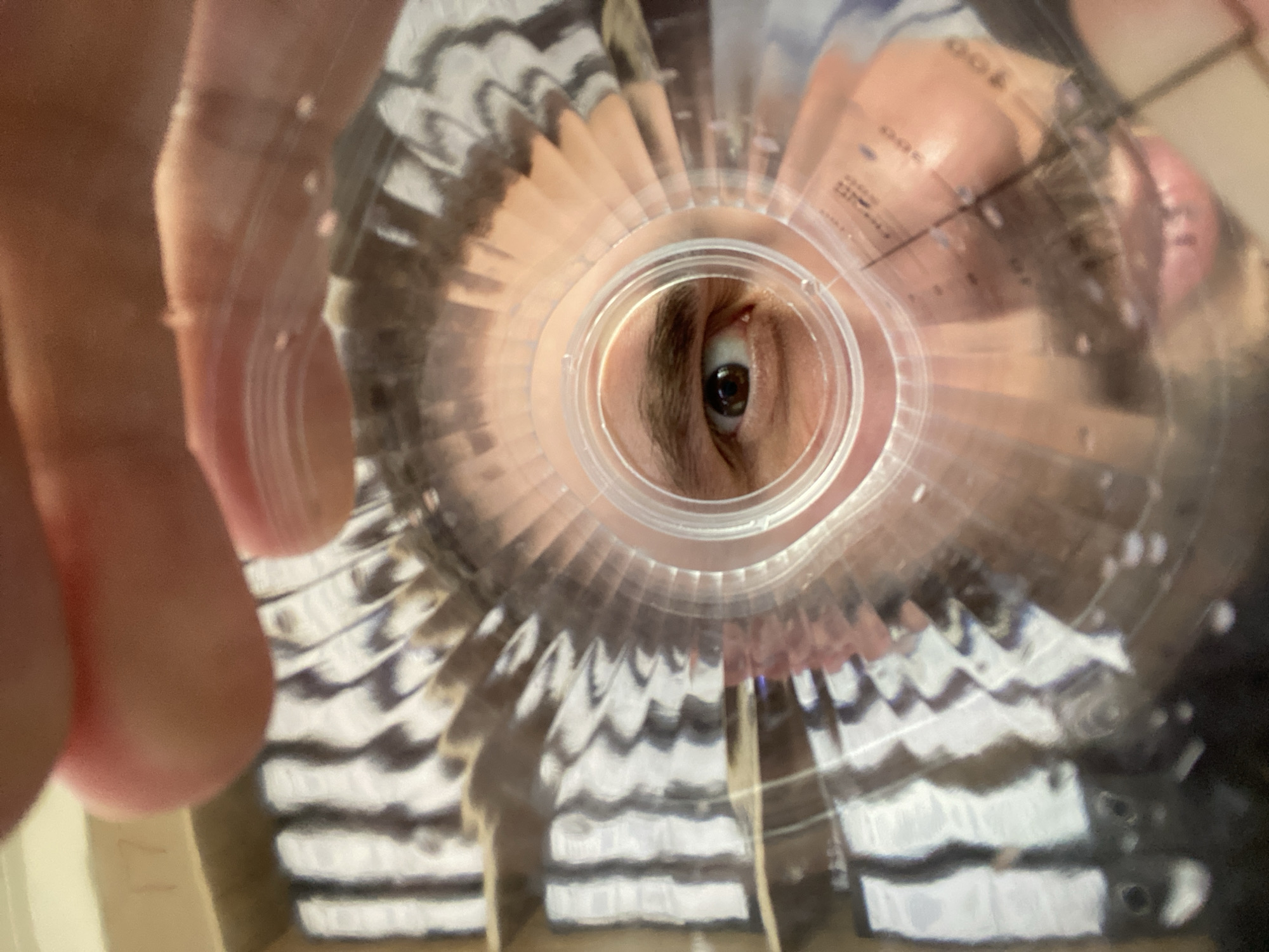 The image is a first-person perspective, looking through the opening of a plastic container. A person's right eye is visible, centered in the opening, with a brown iris and dark pupil. The person's mouth and nose are also partially visible, with pink lips. The container has circular ridges and some markings that appear to be measurements. In the background, out of focus, are shelves containing objects with black and white stripes, possibly books or boxes. The lighting suggests an indoor environment. The location is Luzhitsy, Russia.