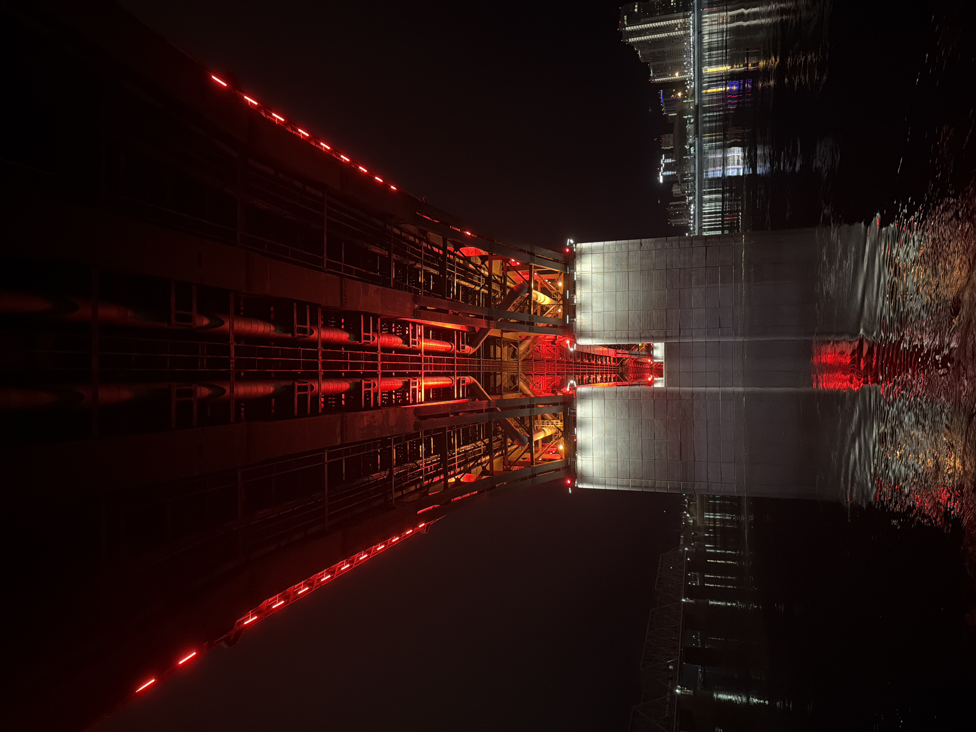 Here's a detailed description of the image:
The image shows the Vinogradovsky Bridge in Krasnoyarsk, Russia, at night, reflected in the Yenisey River. The bridge's metal structure is illuminated with red lights, creating a strong contrast against the dark sky and water. The reflection in the river doubles the effect, creating a symmetrical, almost abstract composition. In the background, city lights and skyscrapers are visible, also reflected in the water, adding depth and context to the scene. The overall atmosphere is dark and moody, emphasizing the industrial feel of the bridge and the urban environment.