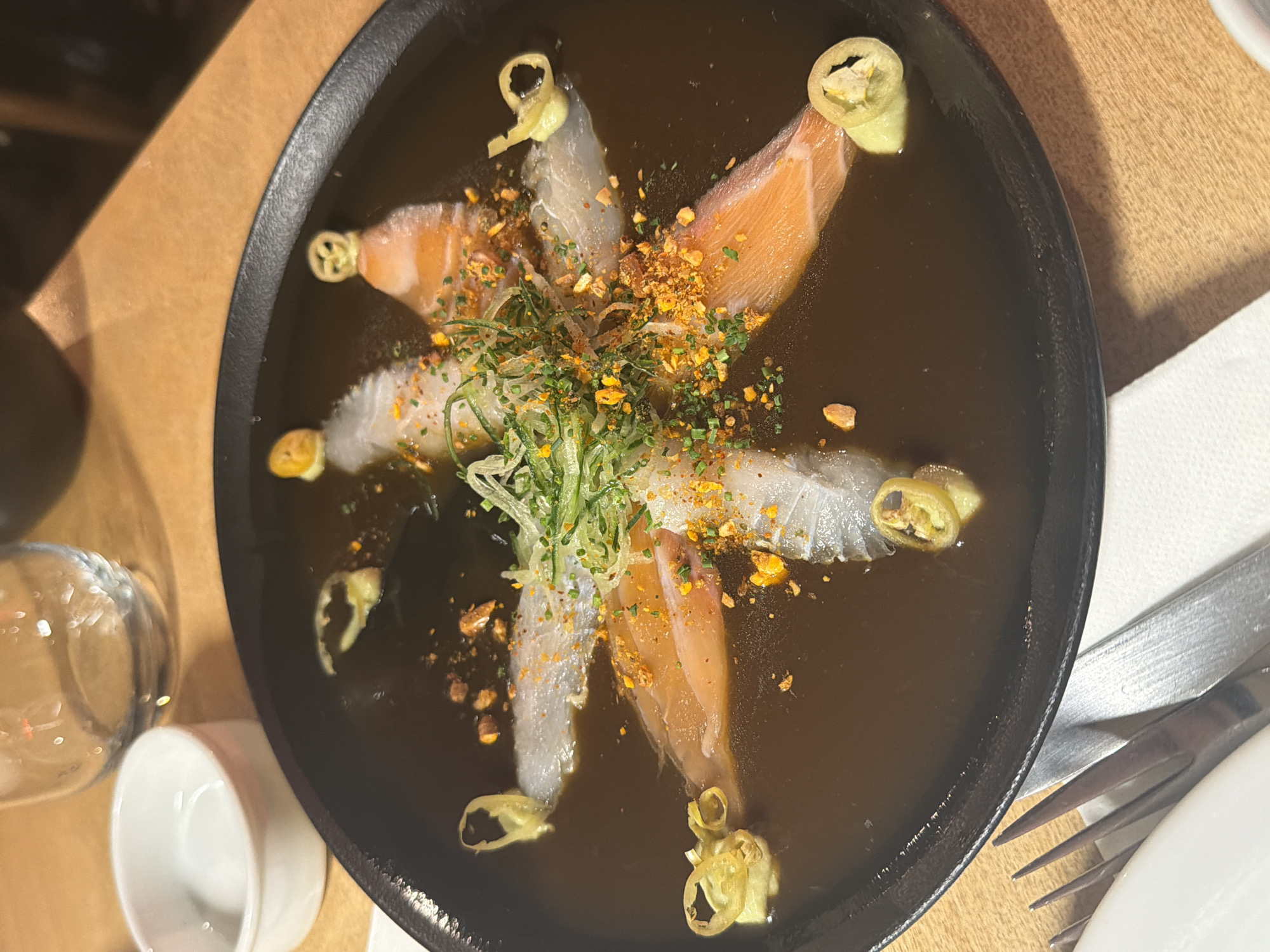 A top-down shot depicts a black, shallow bowl of sashimi in a dark, broth-like liquid. The sashimi consists of six pieces, arranged in a radial pattern, alternating between what appears to be salmon and a lighter-colored fish. Each piece of sashimi is garnished at the tip with a curled, translucent, yellow pepper slice. The center of the arrangement is topped with finely chopped green onions and a yellow, granular seasoning.
The bowl is situated on a light brown, wooden table. To the left of the bowl, there is a clear glass and a small, white porcelain ramekin. To the right, a partially visible white napkin, a silver fork, and the edge of a white plate can be seen. The setting indicates an indoor dining environment.
While the exact location in Buenos Aires, Argentina is not definitively ascertainable from the image, the presence of sashimi, the restaurant setting, and the specific style of food presentation suggest a contemporary or fusion-style dining establishment in an urban area.