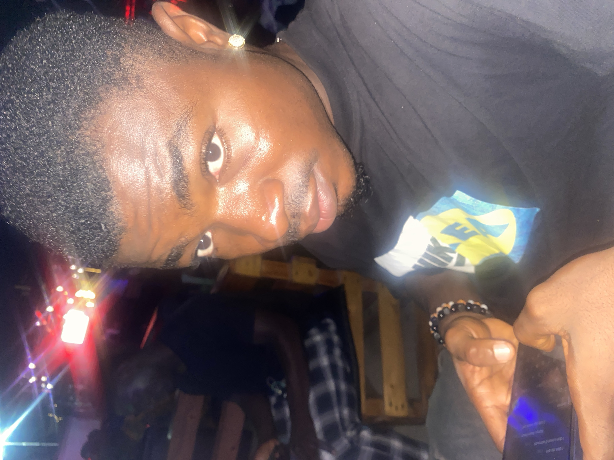 A dark-skinned male is prominently featured, gazing forward with direct eye contact. He has short, dark hair with faded sides, faint facial hair comprising a mustache and short beard, and a single gold stud earring in his right ear. He wears a black t-shirt with a partially visible blue and yellow graphic on the chest and a multi-colored beaded bracelet on his left wrist. Both hands hold a dark-colored smartphone, which emits a blue light from its screen. In the blurred background, another male in a dark t-shirt and plaid trousers is seated on wooden furniture. The scene is dimly lit by multiple artificial light sources of varying colors (red, white, yellow) originating from the distant background, indicative of a casual, possibly outdoor or semi-outdoor, venue in Howayi, Nigeria, furnished with what appears to be wooden benches or pallet-style seating.