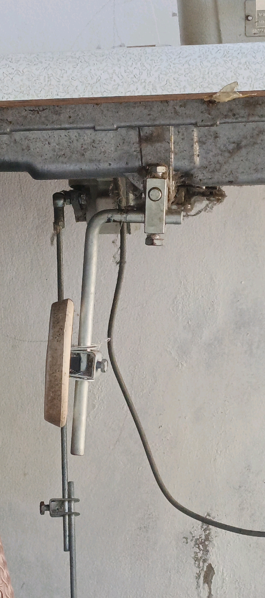 The image shows a close-up view of some plumbing and wiring attached to a metal surface, possibly part of a machine or a building fixture. The plumbing consists of several pipes, some of which appear to be made of galvanized steel, connected with various fittings and brackets. There is also a thick, dark electrical wire that runs diagonally across the frame. A wooden or plastic object, rectangular in shape, is attached to one of the vertical pipes. The overall scene appears to be somewhat aged and possibly neglected, with some dust and debris visible on the metal surfaces. The background is a textured, light-colored wall. The location is identified as Sukawati, Indonesia. No people are visible in the image.