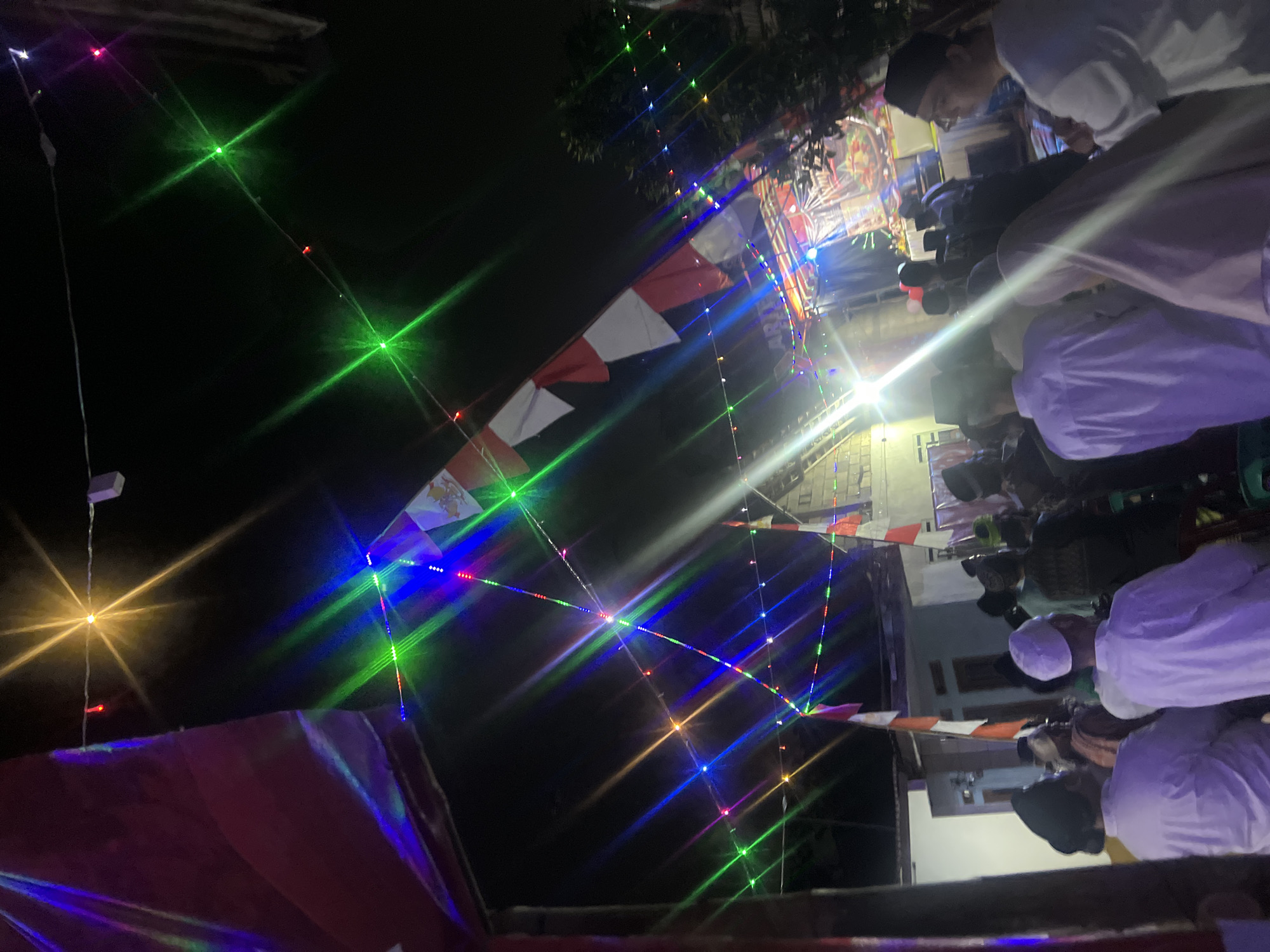 The image depicts an outdoor nighttime gathering, likely a cultural or religious event, in Klapanunggal, Indonesia. A crowd of people, predominantly men dressed in white attire and skullcaps, are seated and standing, facing a stage area illuminated by colorful string lights and stage lighting. The lights create a vibrant, festive atmosphere with prominent light flares. Indonesian flags, red and white, are visible, suggesting a national celebration or a significant local event. Tents or awnings are set up, providing some overhead cover. The background reveals simple buildings and some foliage, indicating a semi-rural or residential setting within the city. The overall scene conveys a sense of community and shared observance.