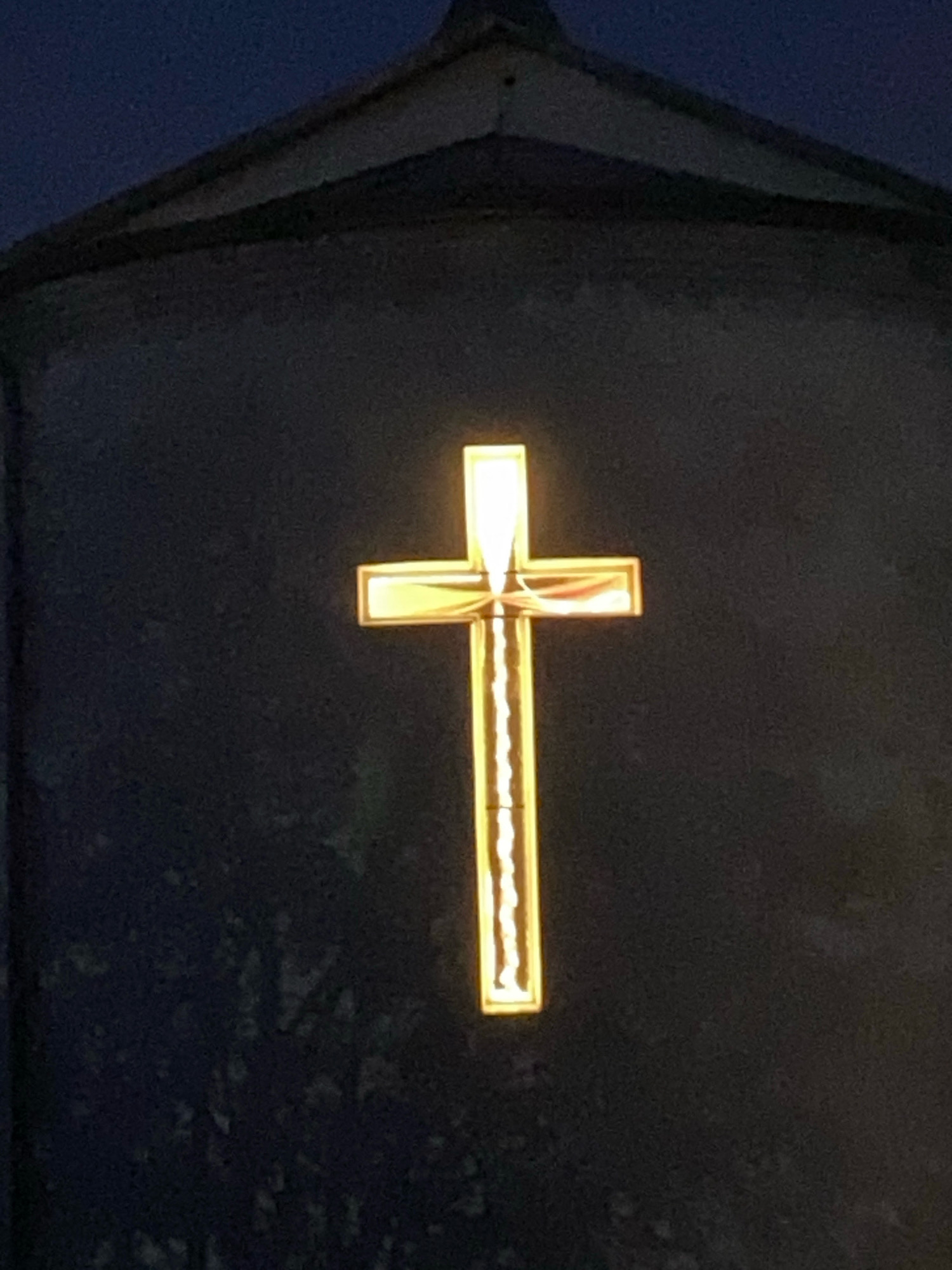 The image shows a close-up of a lit, gold-colored cross against a dark background, possibly a building facade. The cross is illuminated from within, with a distinct segmented light source visible along its length. The top of a dark-colored, peaked roof structure is visible above the cross. The lighting suggests the photograph was taken at dusk or night. This cross is likely located on the facade of the Church of the Exaltation of the Holy Cross in Machulishchy, Belarus, a prominent landmark in the city.