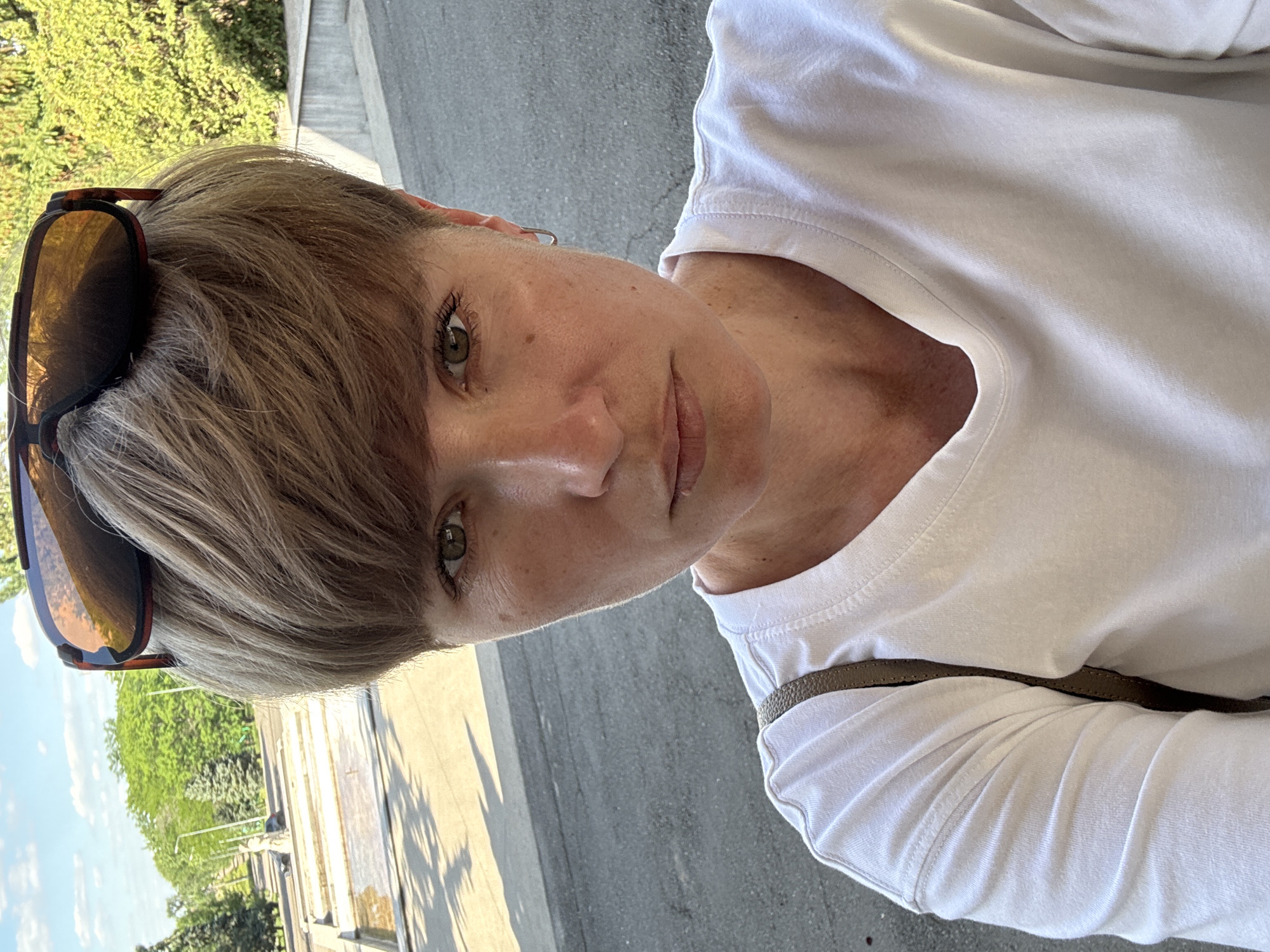 An outdoor selfie shows a woman from the chest up, facing the camera. She has short, light brown hair styled with bangs, and wears sunglasses perched on top of her head. Her eyes are green, and her expression is neutral. She is wearing a white t-shirt and a brown purse strap is visible over her left shoulder. The background appears to be an urban setting with trees, a paved area, and a structure with multiple levels. The lighting suggests it is daytime.