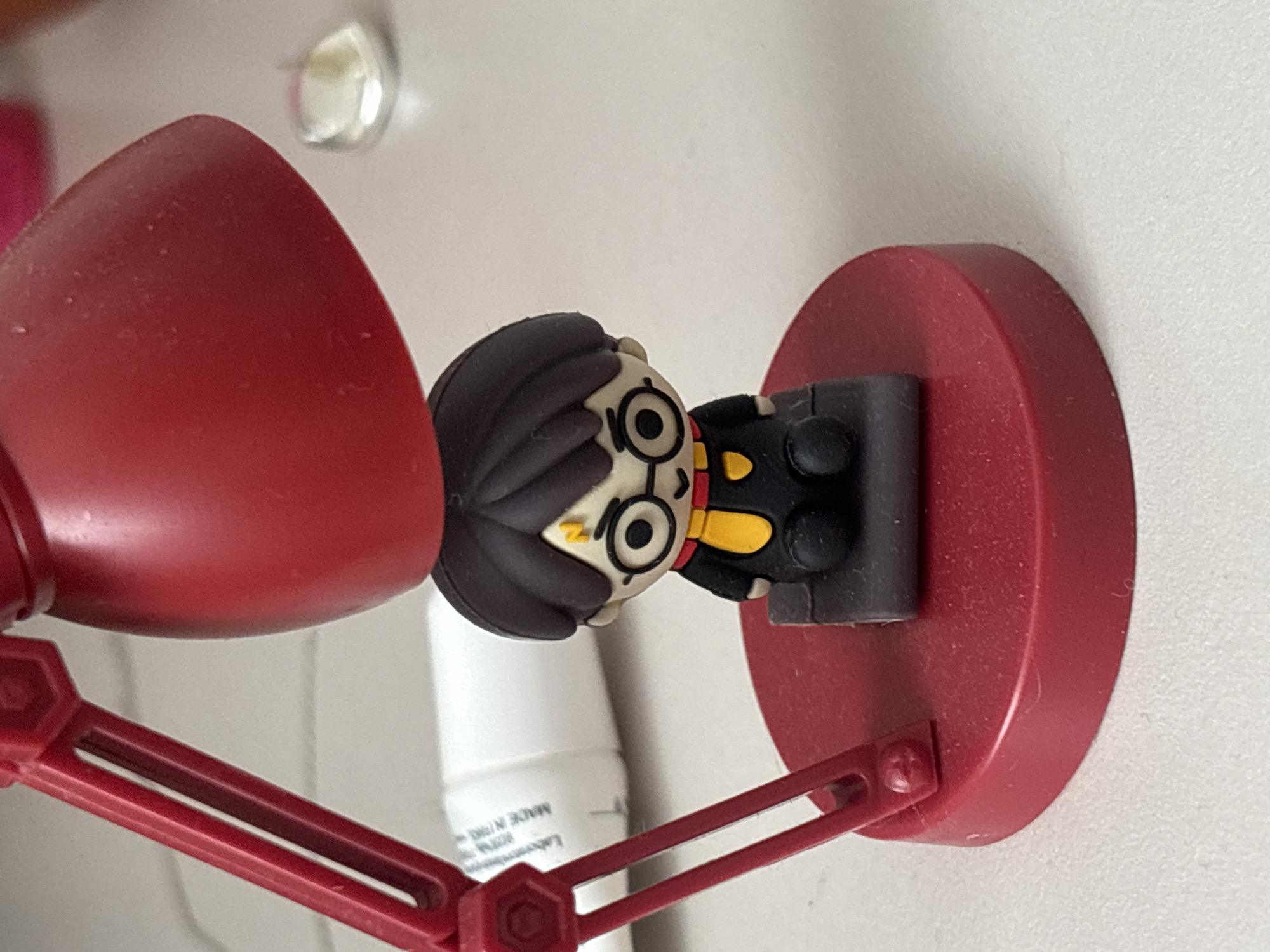 A low-angle, close-up shot depicts a red desk lamp affixed to a white wall. A Harry Potter figurine with black hair and round glasses is positioned adjacent to the lamp's shade. The lamp has a hexagonal joint in its arm and a circular base. A white tube is visible in the background. The location is unspecified.