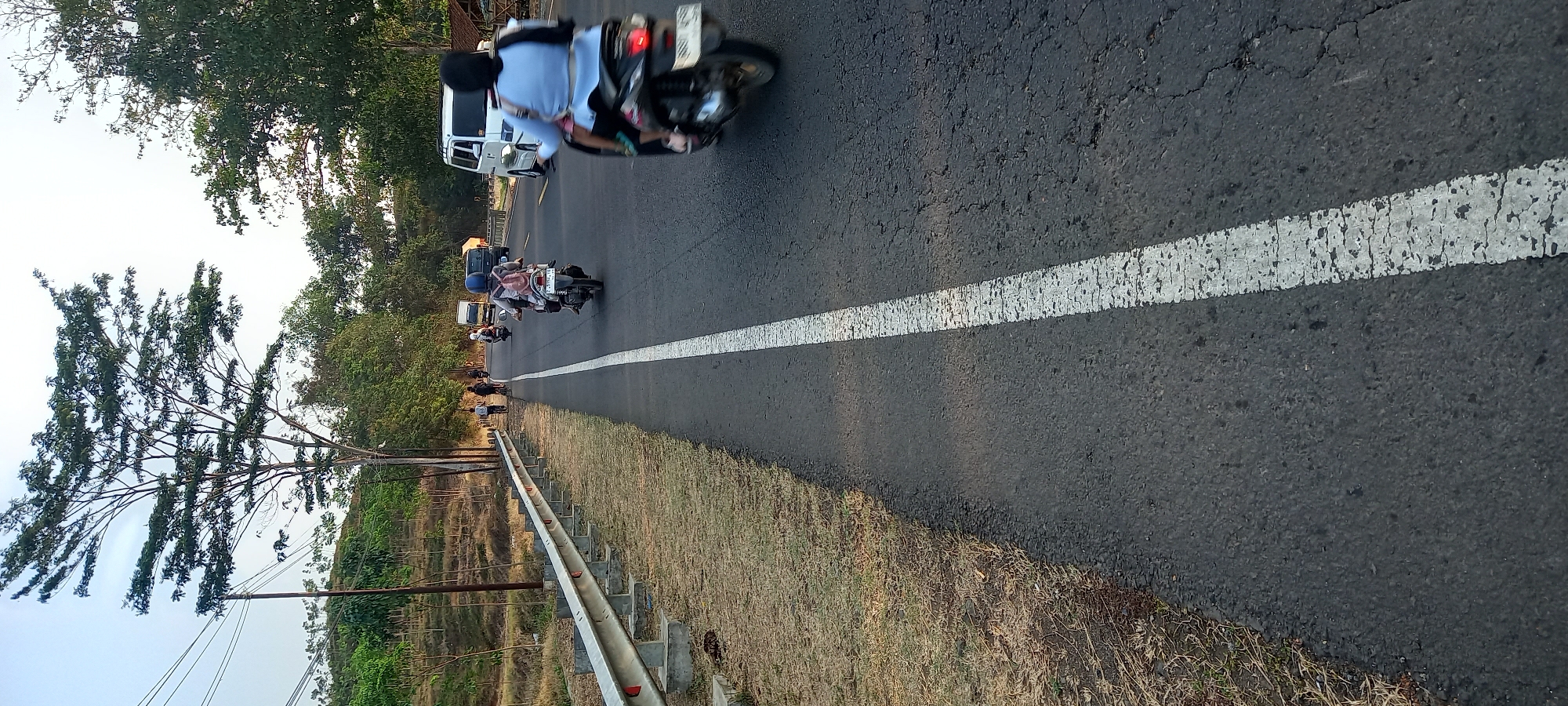 In Adimulya, Indonesia, a rural road stretches out under a hazy sky, bordered by dry grass and a metallic guardrail on the left. The asphalt road is marked by a prominent white line, cracked and weathered. Several motorbikes and a few bicycles traverse the road, with riders visible. In the background, trees line the horizon, and a white van and truck are present. The lighting suggests it's late afternoon.