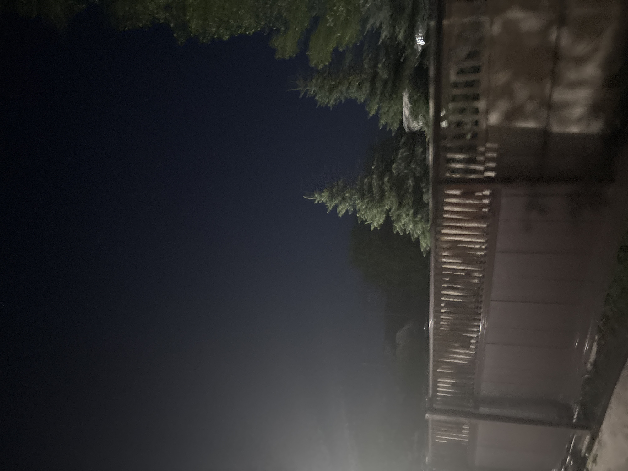 The image shows a dark, outdoor scene at night, with a wooden fence in the foreground. Behind the fence, several evergreen trees are visible, with the sky above them appearing dark blue or black. There is some artificial light source causing a bright, hazy area in the upper left quadrant of the image. The fence is made of vertical wooden planks and has a horizontal railing at the top. The overall impression is of a residential backyard or a similar private outdoor space. The location in the city of Orekhovka, Russia is not discernible from the image itself.